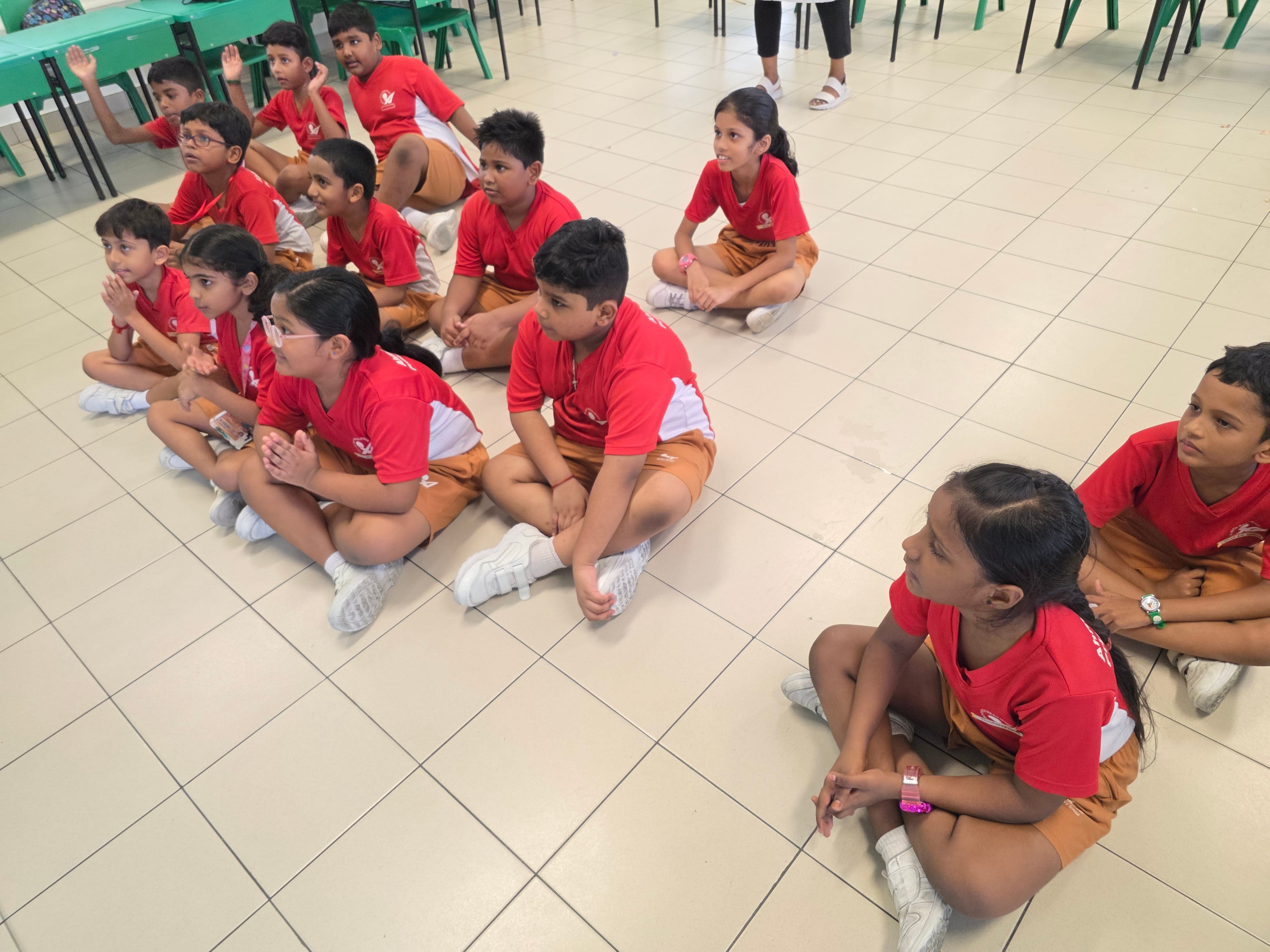 A group of young school children in red and tan uniforms sitting cross-legged on a tiled floor indoors.