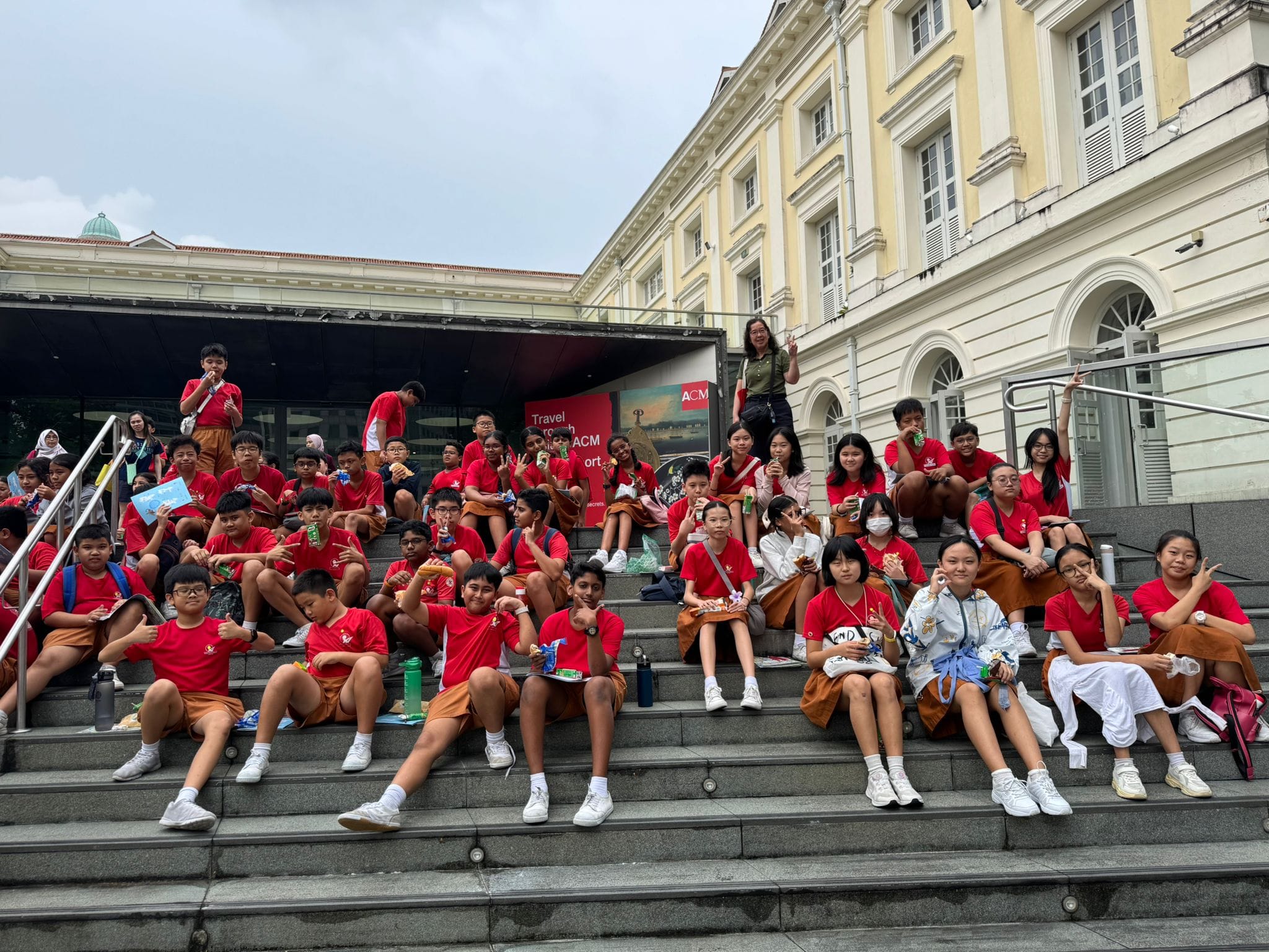 A large group of students in red uniforms sitting on outdoor steps in front of a colonial-style building.
