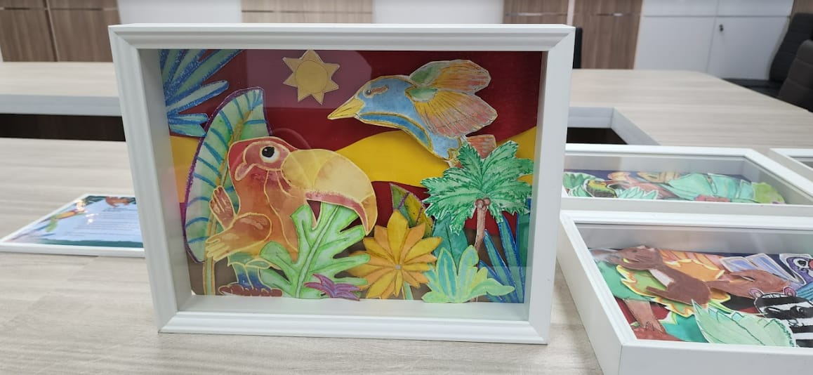 White framed shadow box with colorful paper-cut tropical scene featuring a toucan, flying bird, sun, and palm trees.