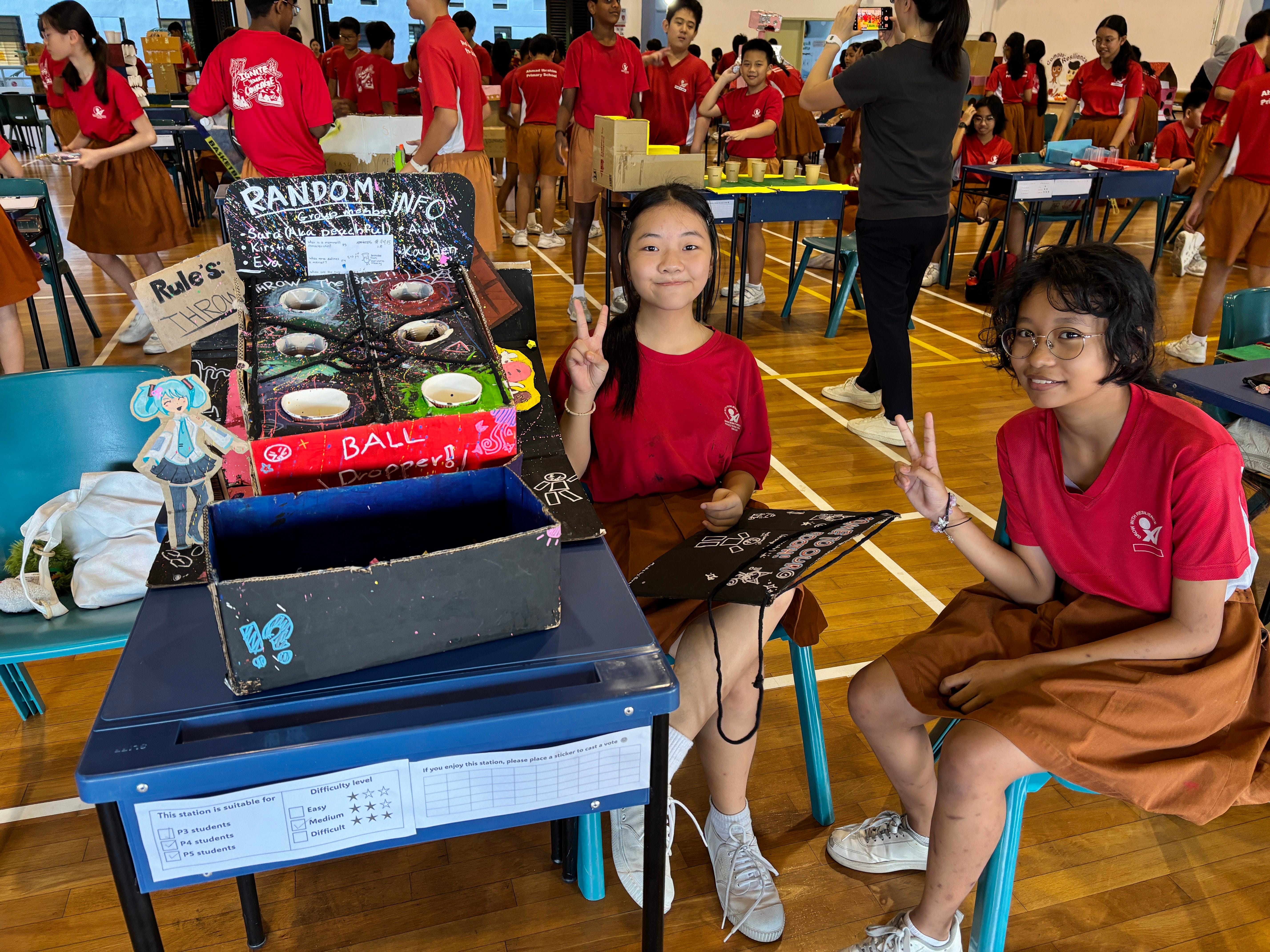 Two girls in red uniforms pose with peace signs beside a handmade Ball Dropper game at a school fair.