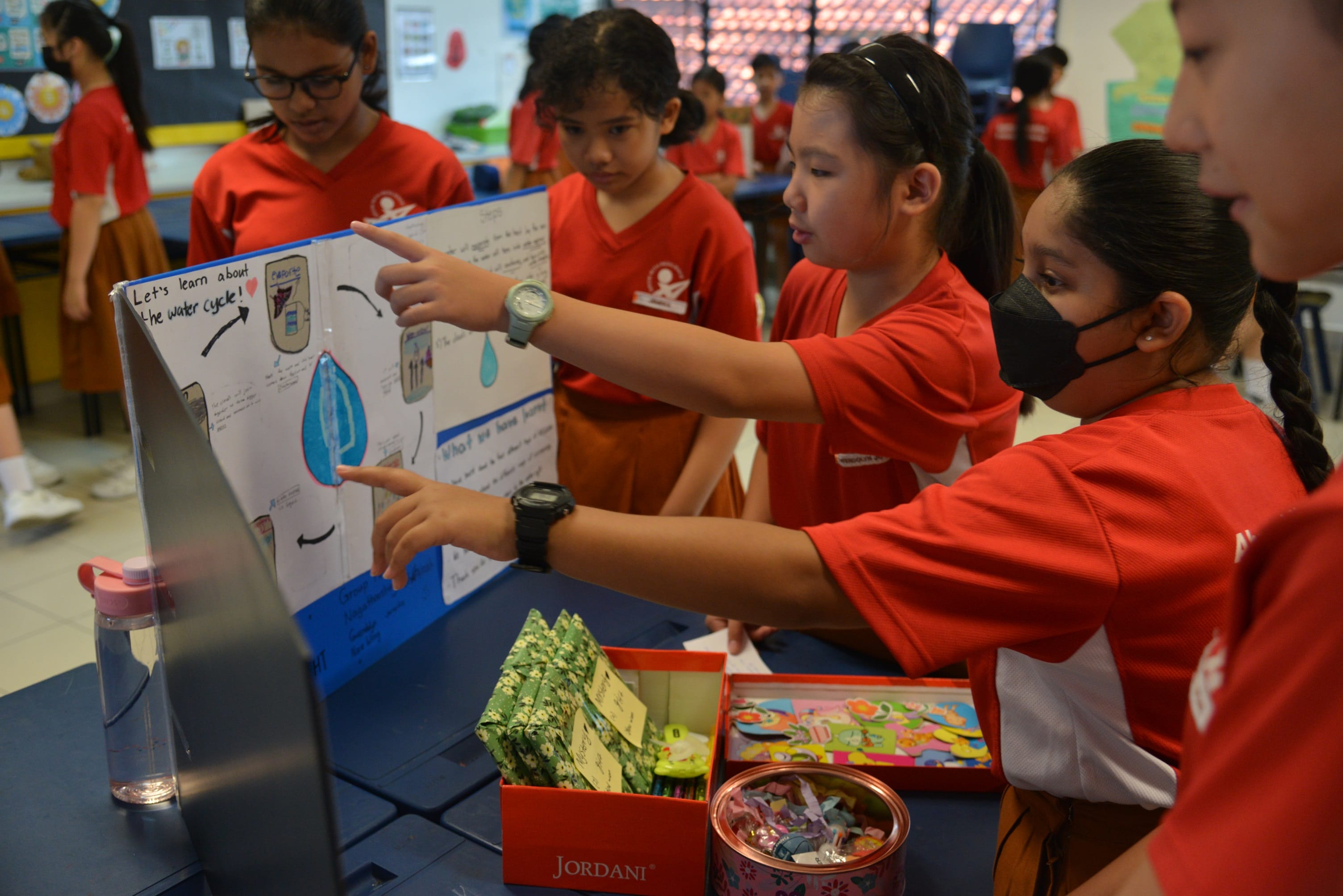 Students in red uniforms point at a handmade poster about the water cycle during a school presentation.