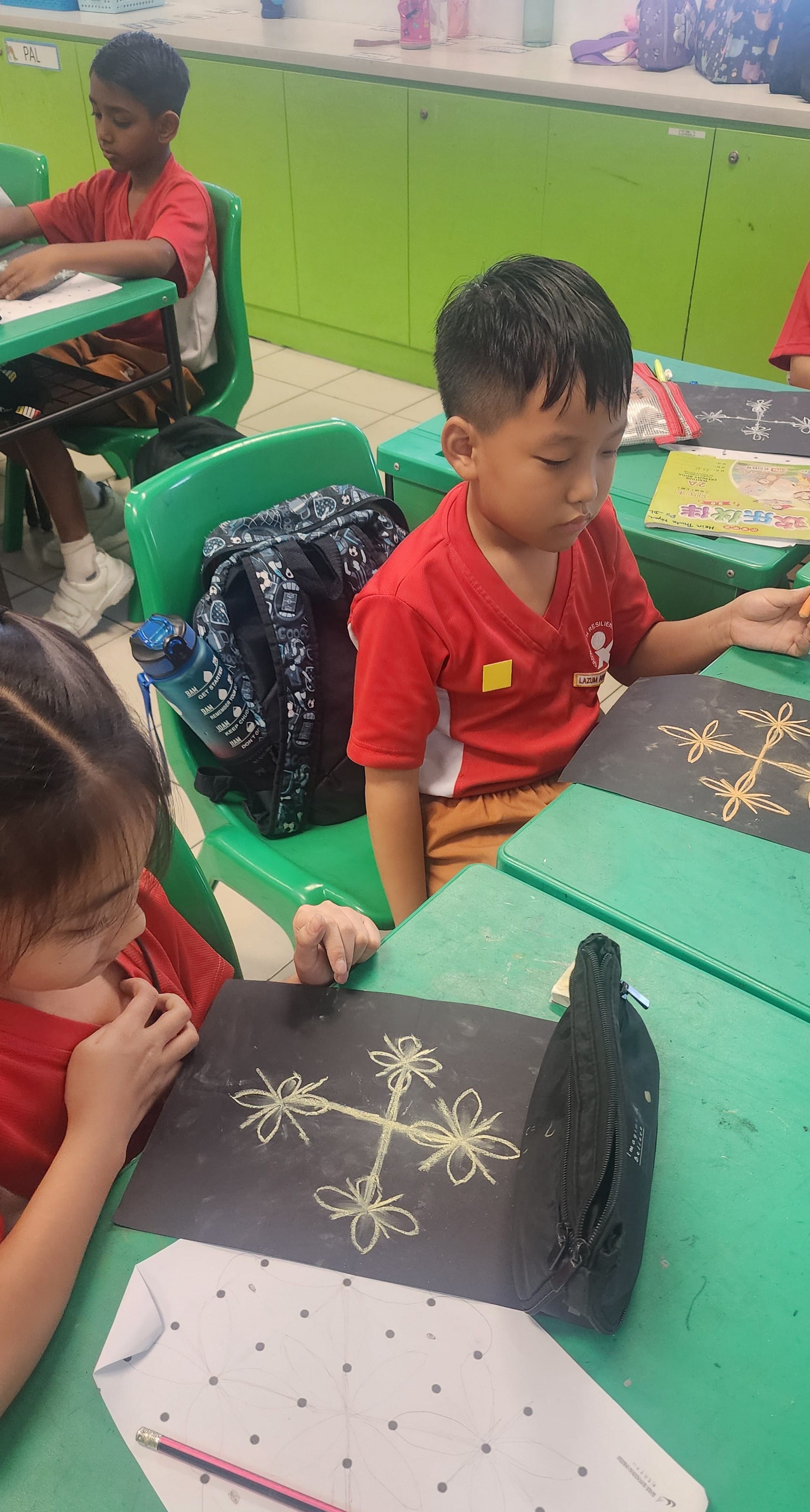 Young students in red uniforms sit at green desks, drawing flower patterns on black paper with gold lines.