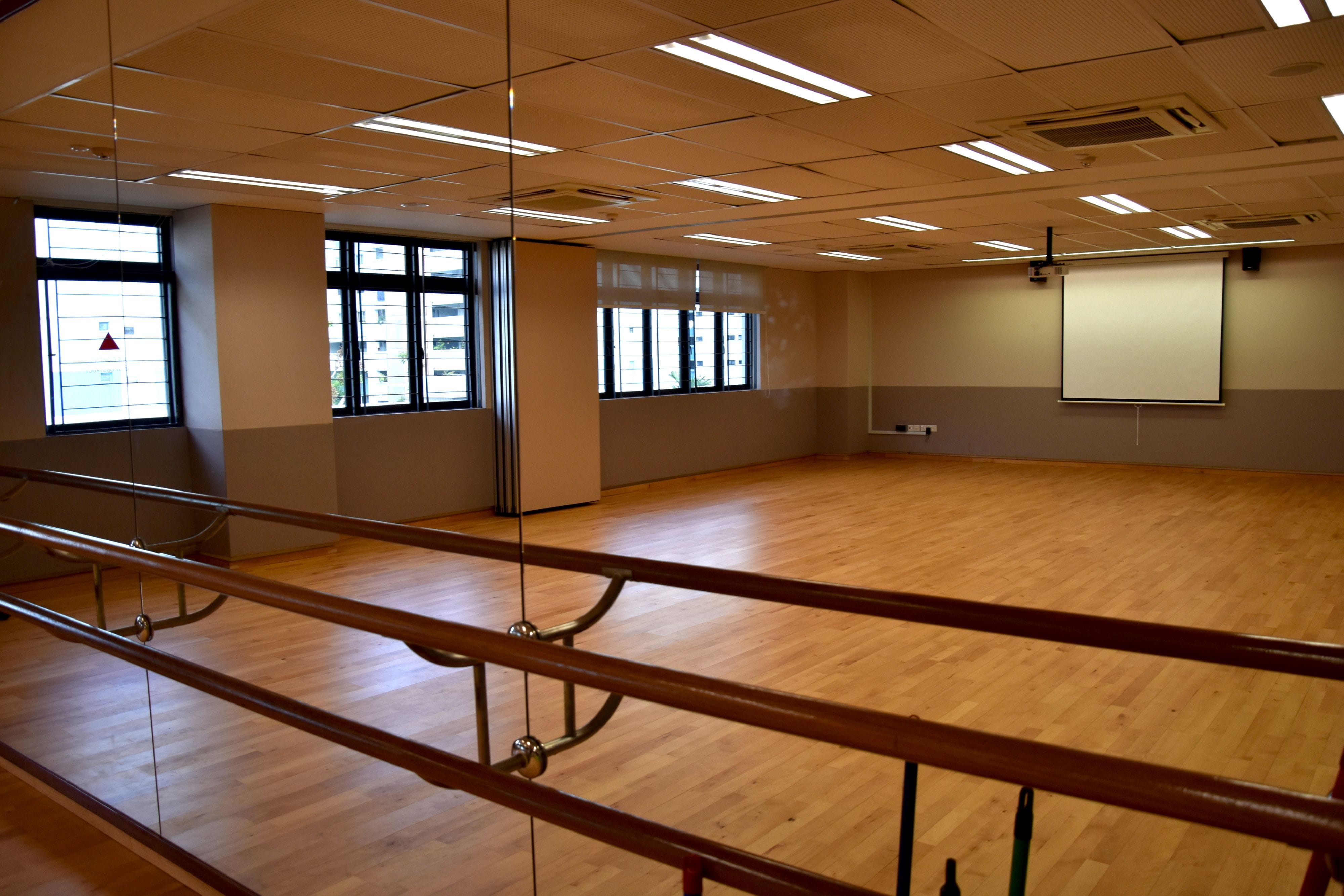 Empty dance studio with wooden floor, wall mirrors, ballet barres, large windows, and a projector screen on the wall.