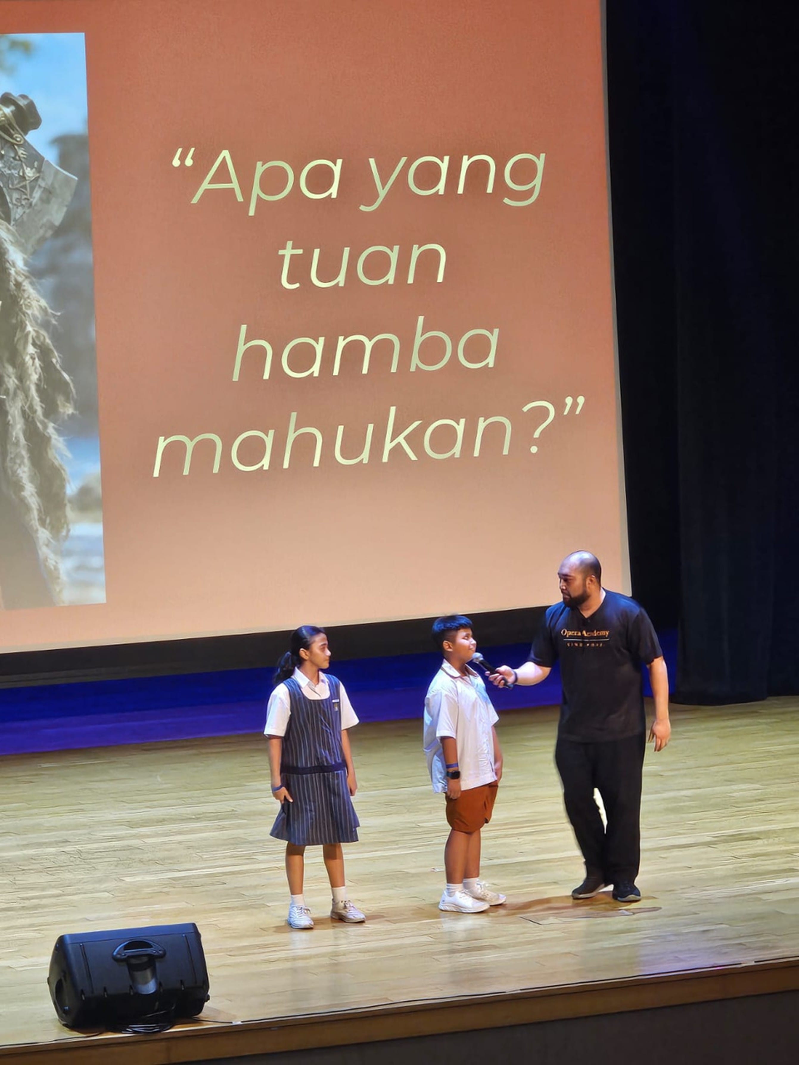 An adult holds a microphone to a child on stage beside another child, with a Malay text slide projected behind them.