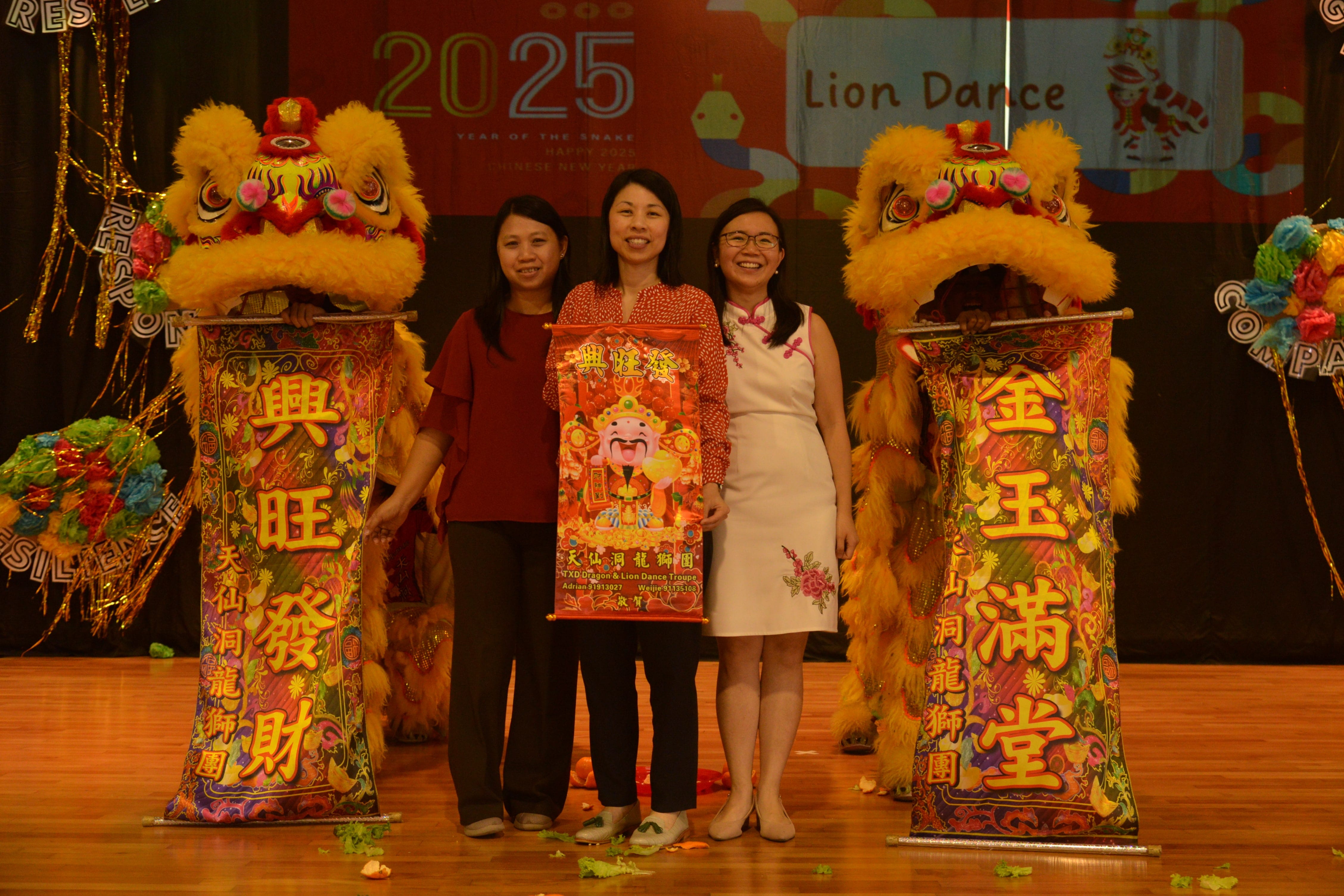 Three women pose on stage holding a red scroll between two lion dance costumes at a 2025 Chinese New Year event.