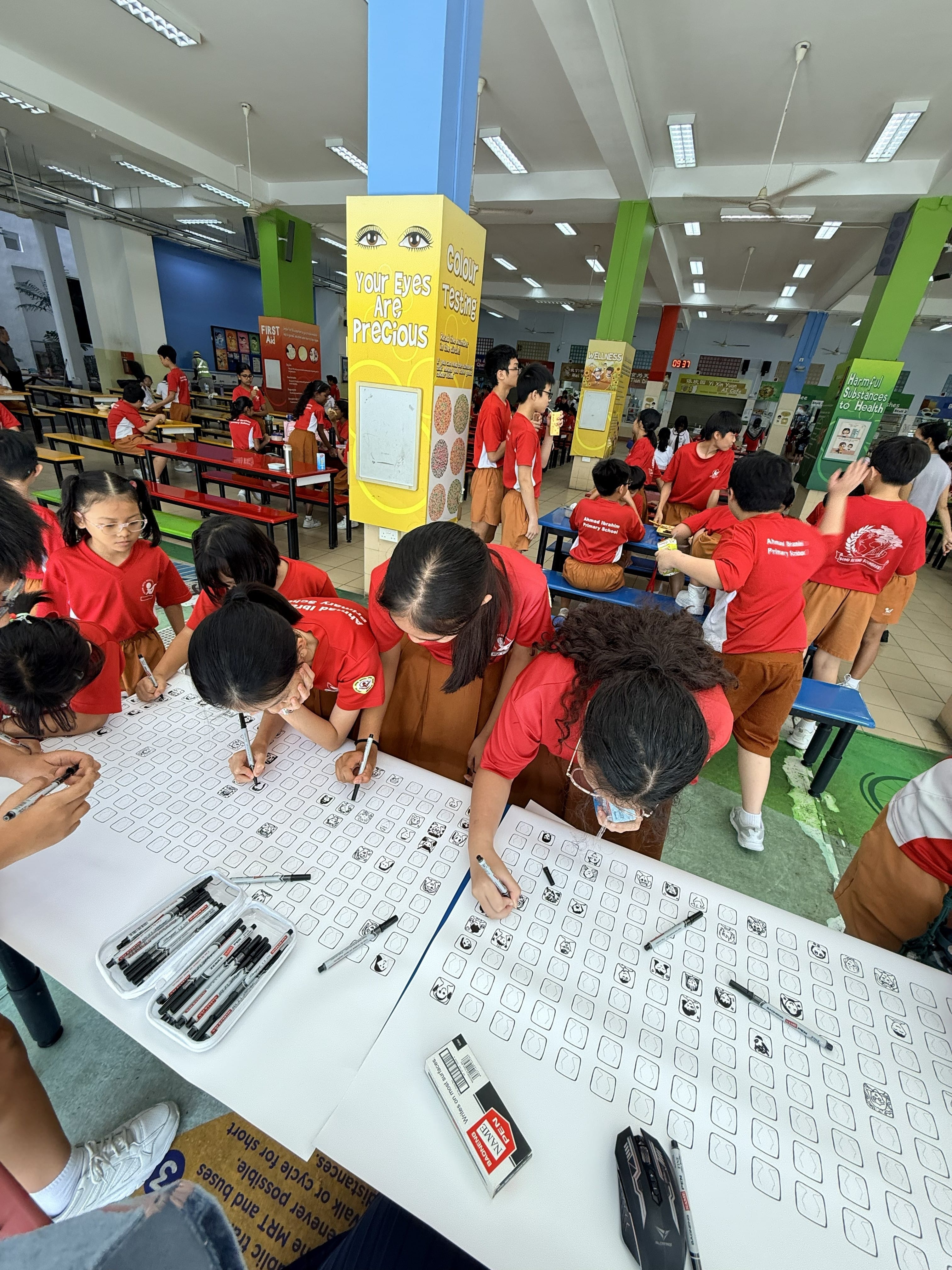 Students in red uniforms draw with markers on large sheets at tables in a school hall with health education displays.