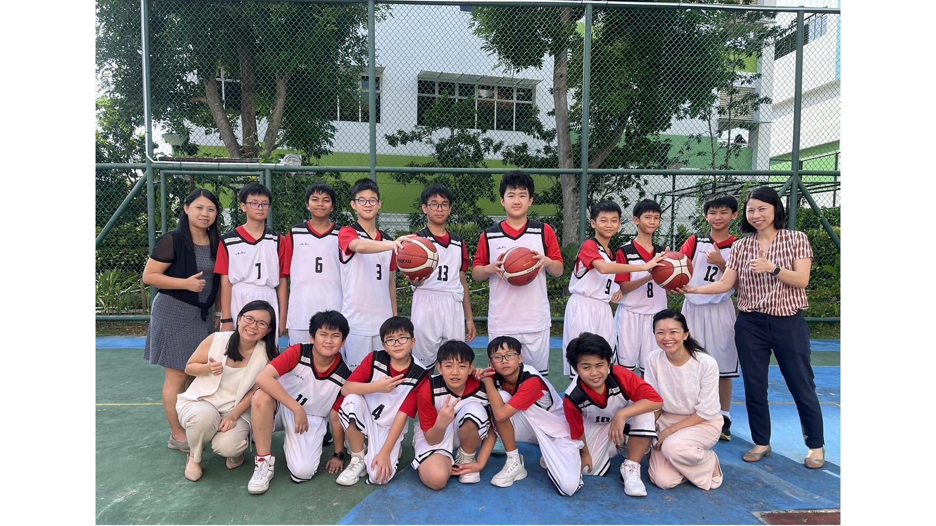 A youth basketball team in white and red uniforms poses with basketballs on an outdoor court with two adult women.