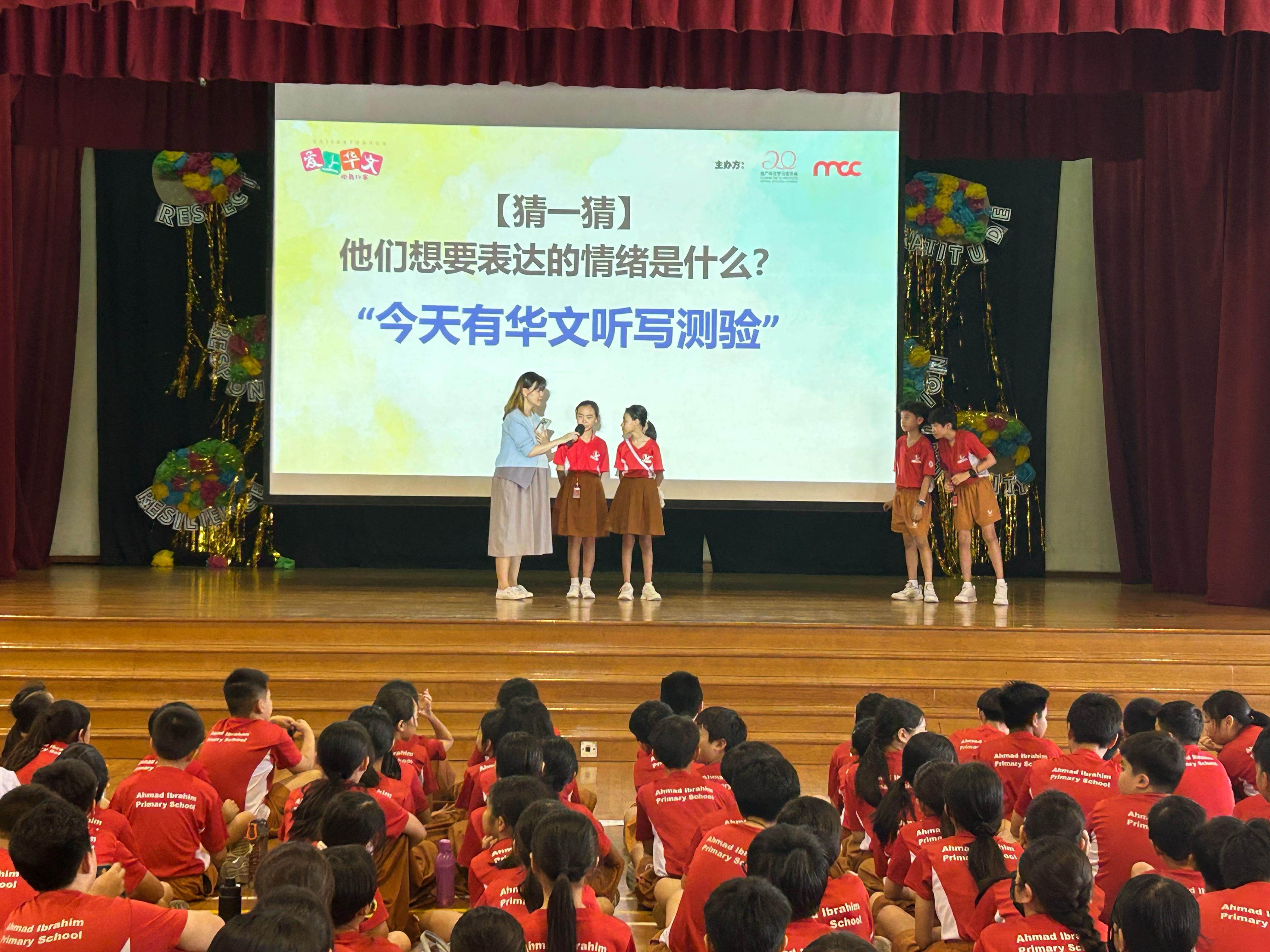A teacher holds a microphone for two students on stage at Ahmad Ibrahim Primary School with a Chinese text slideshow.