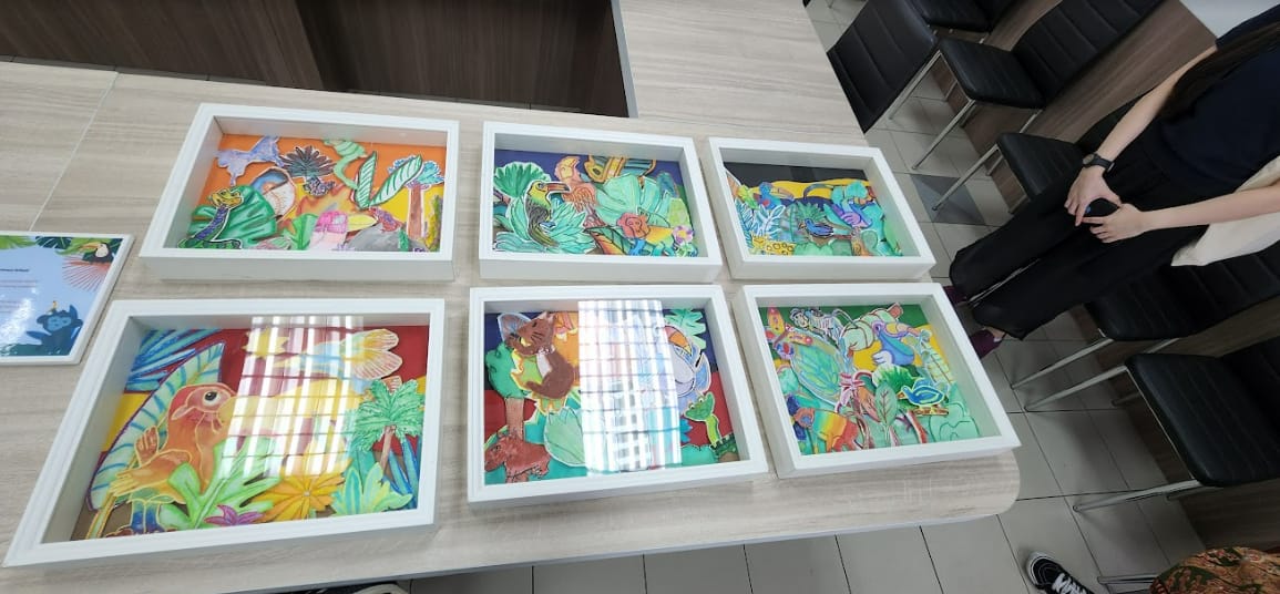 Six colorful framed artworks with tropical animals and plants displayed on a wooden table, with a person standing nearby.