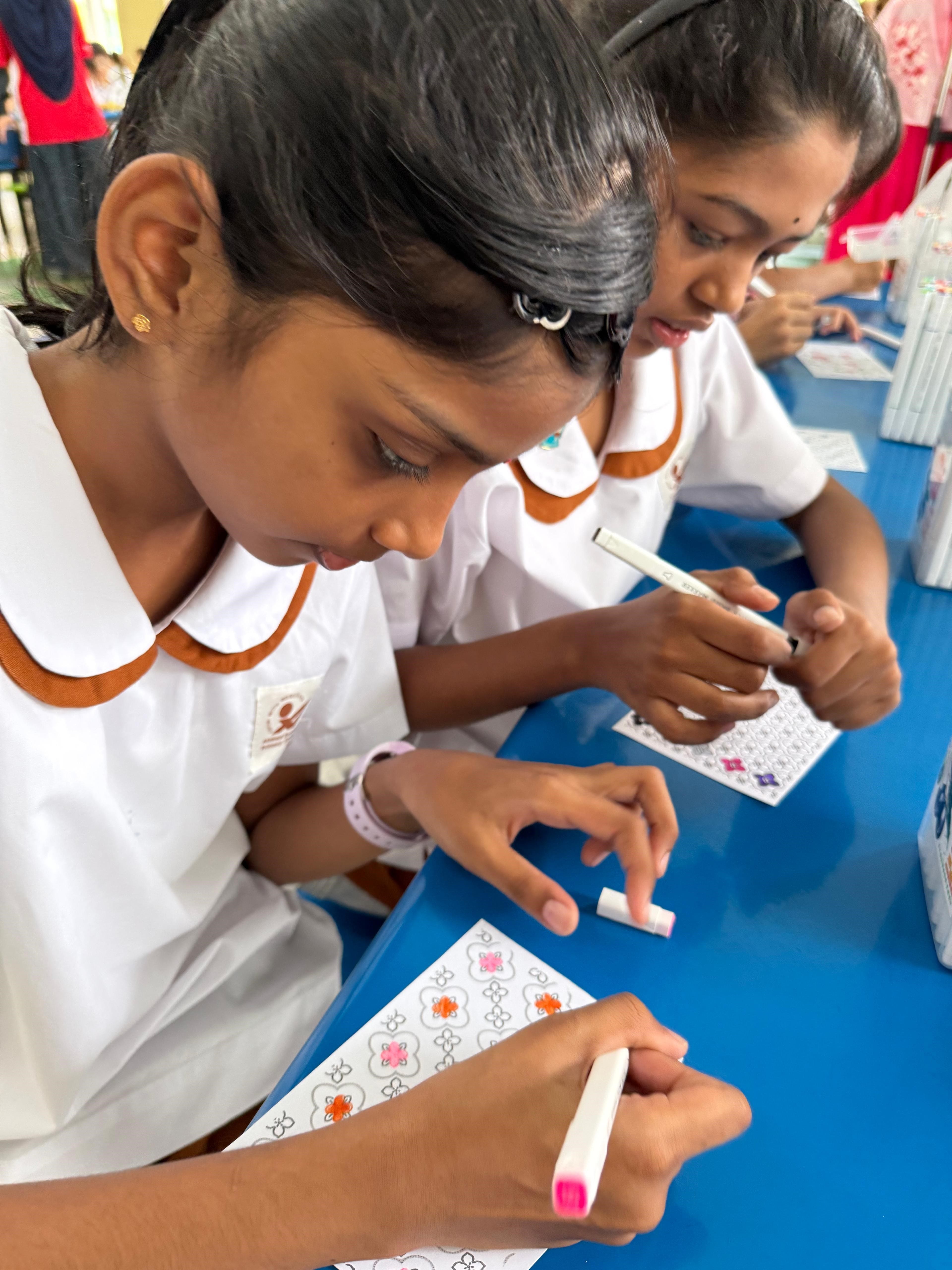 Two girls in white school uniforms color patterned cards with markers at a blue table.