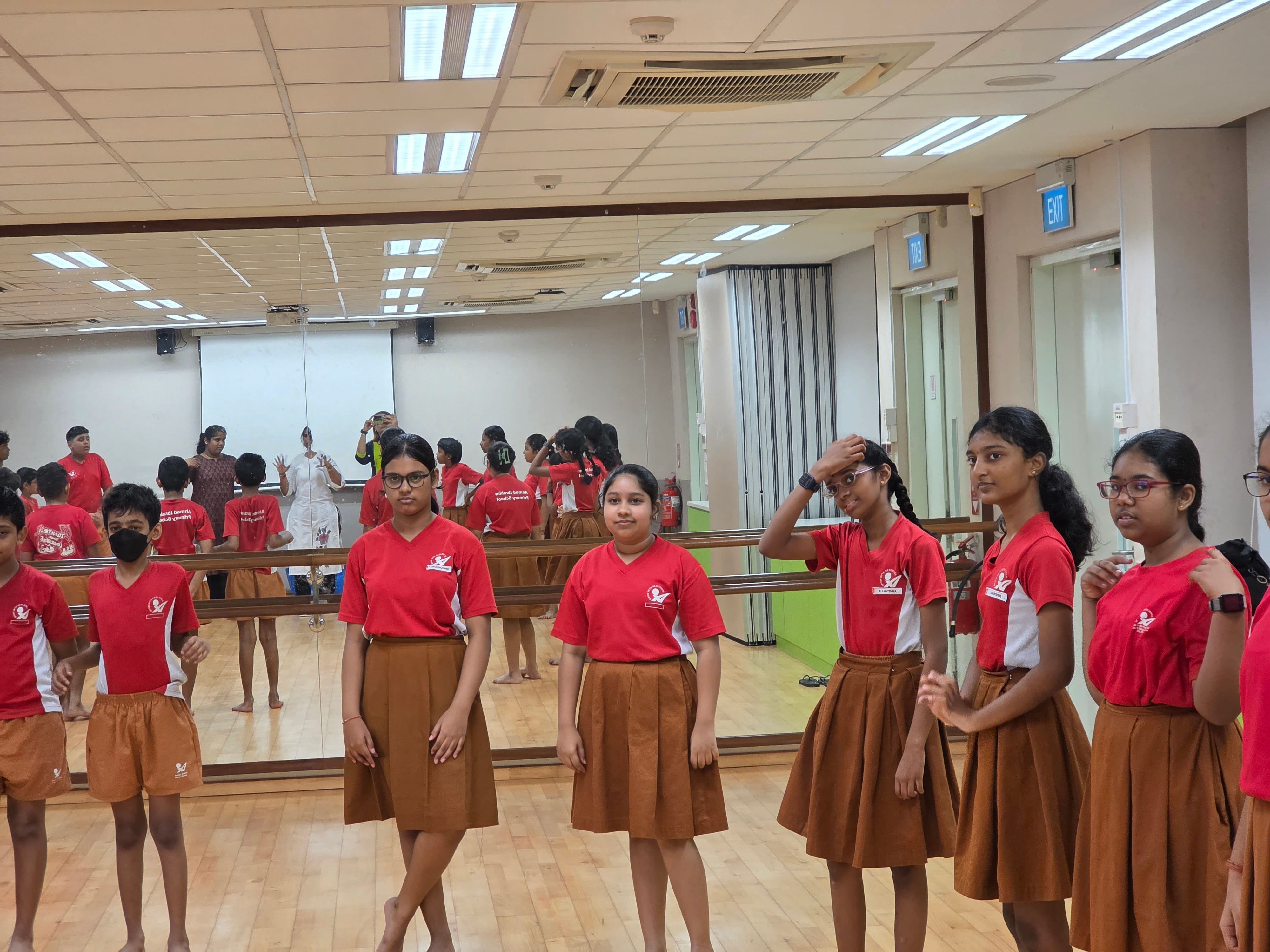 Students in red shirts and brown skirts stand in a mirrored dance studio with a teacher visible in the reflection.
