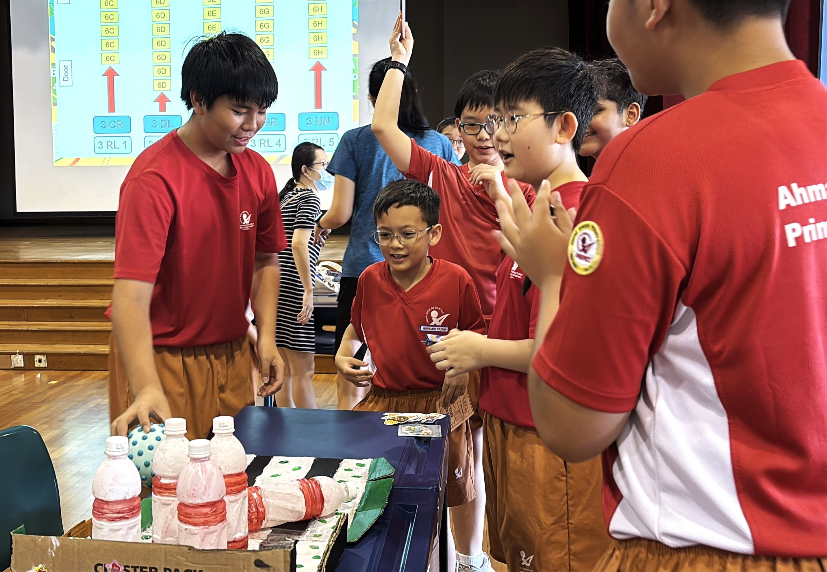 Several primary school students in red uniforms gathered around a table with a cardboard craft project made from plastic bottles.