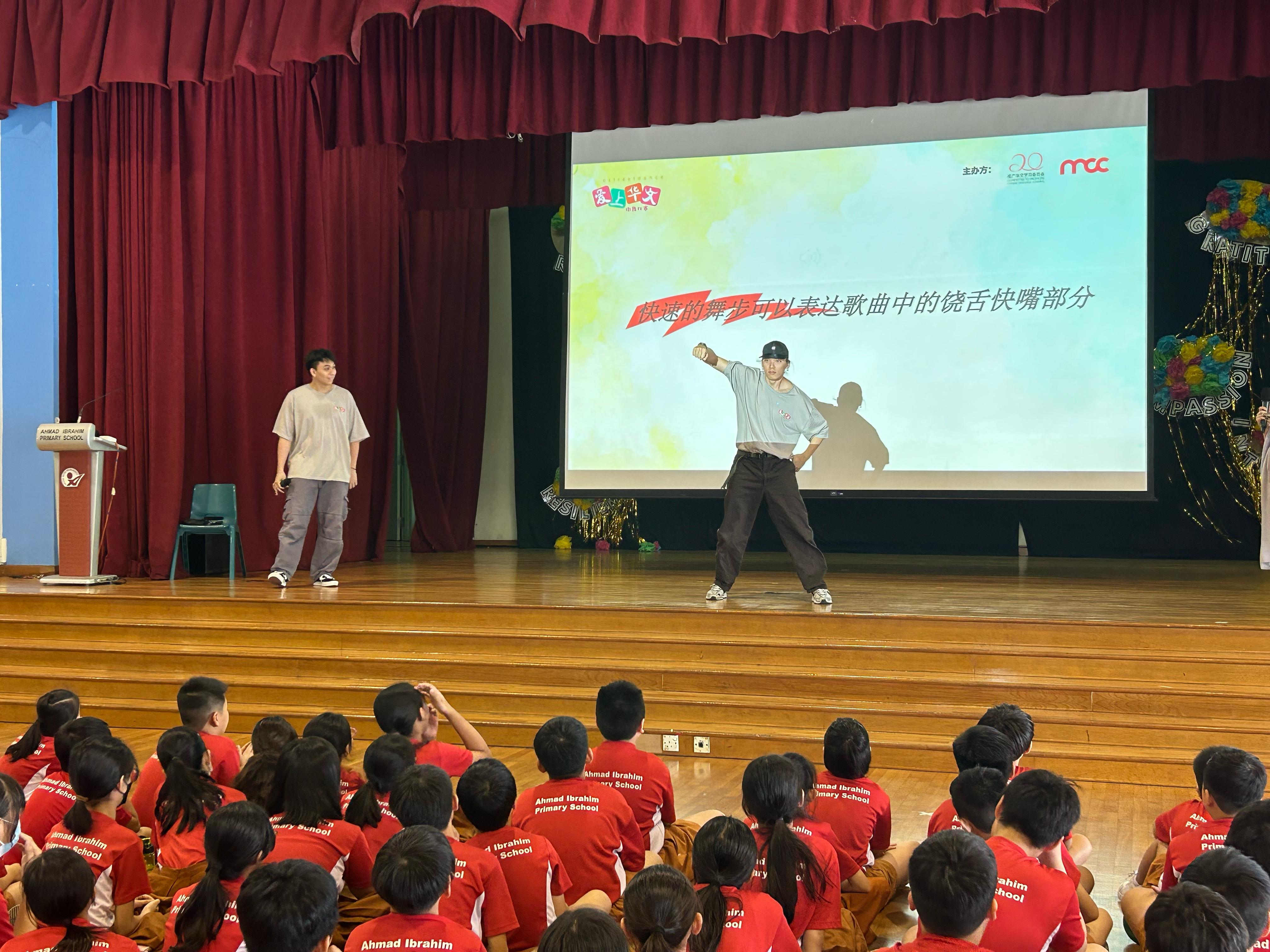 Two performers dance on a school stage before students in red uniforms, with a Chinese-text projection behind them.