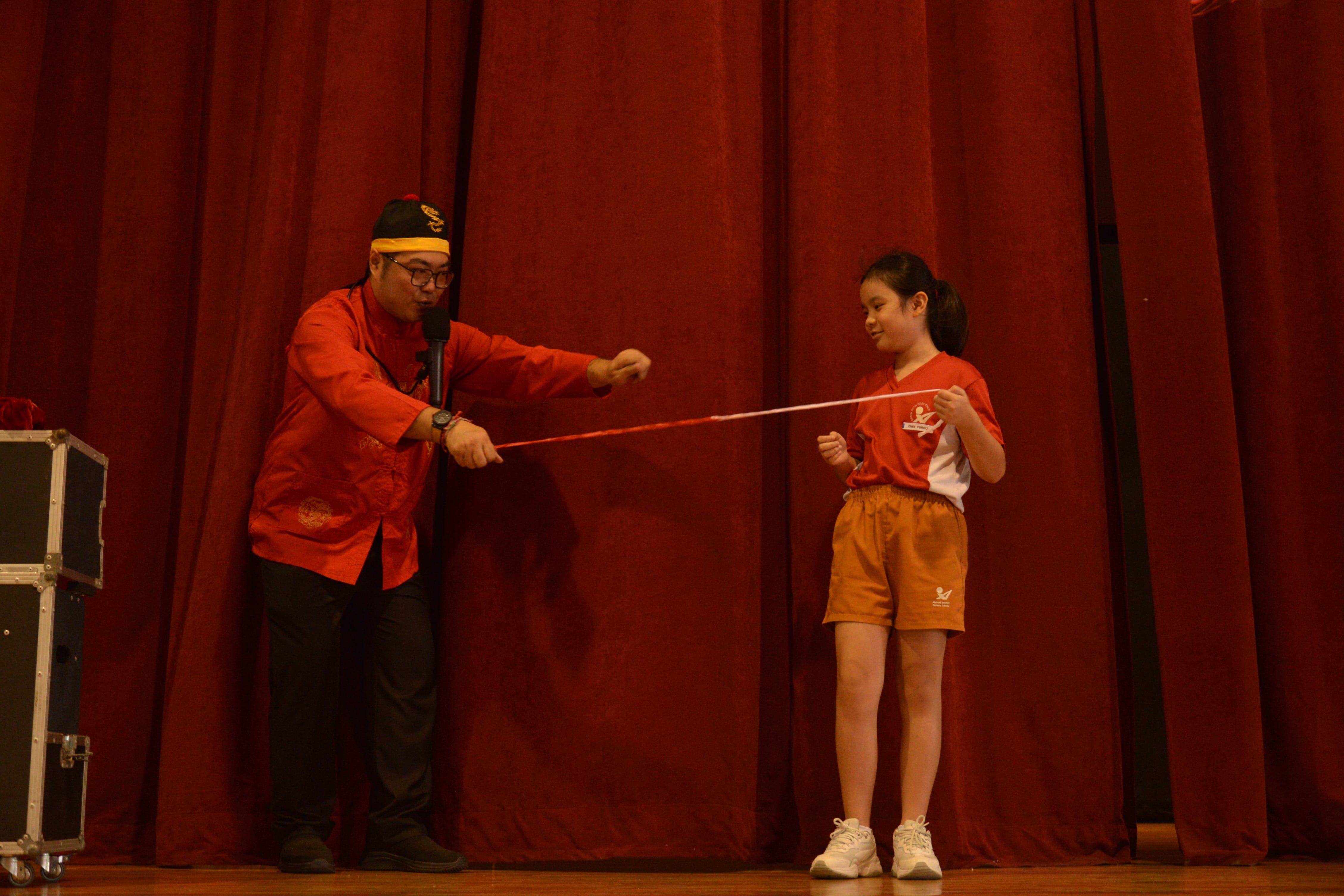 A man in a red traditional jacket holds a rope with a young girl on a stage with red curtains behind them.