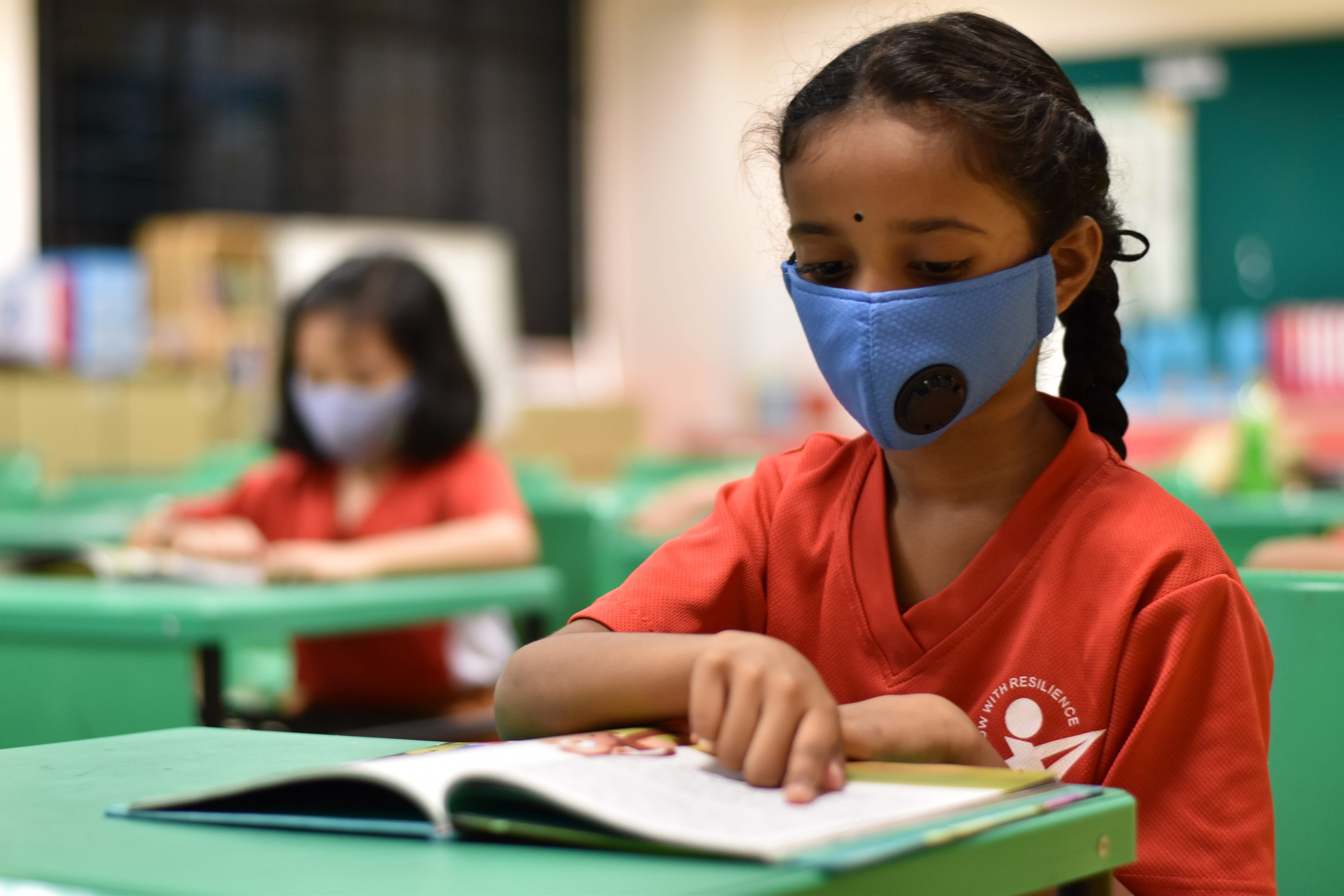 Two girls wearing face masks sit at green desks in a classroom, reading open books.