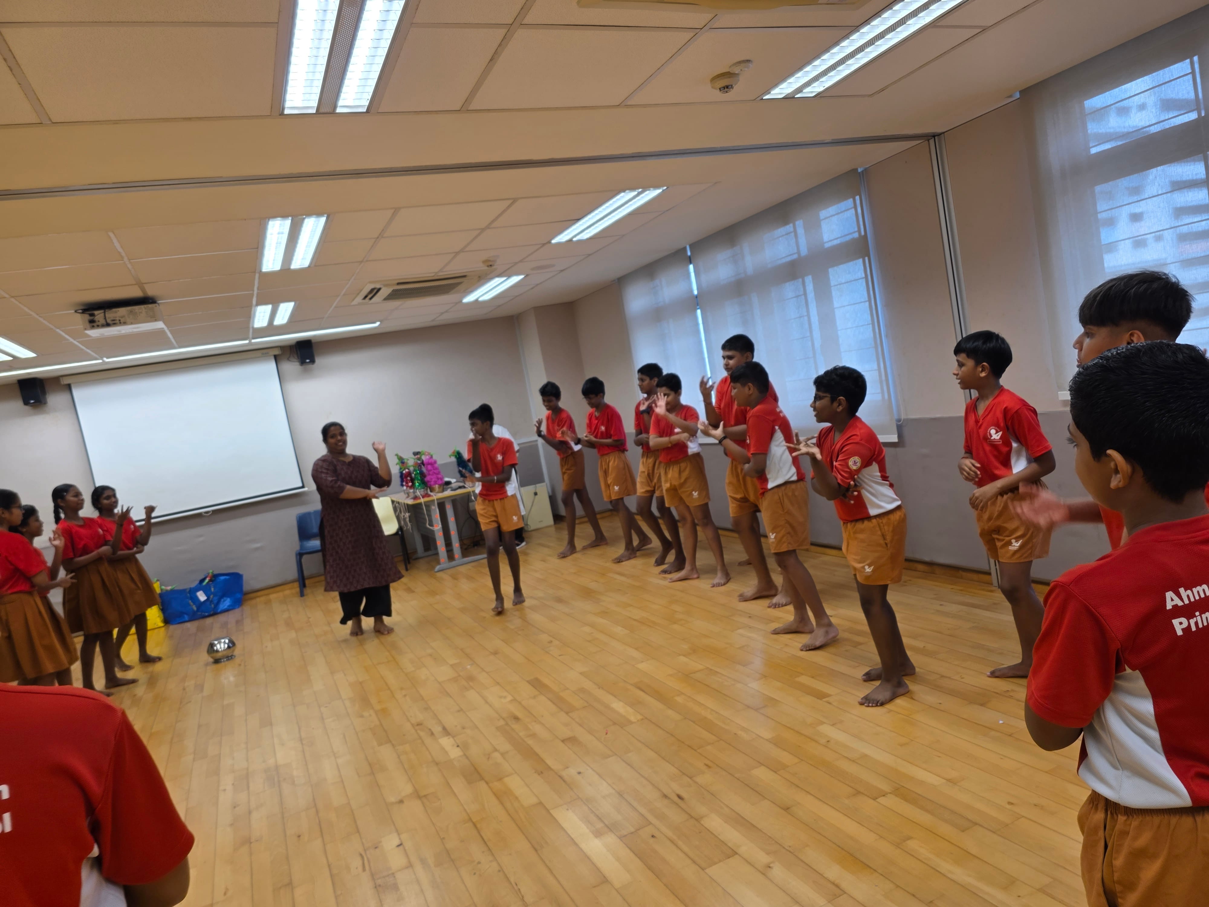 Children in red shirts and tan shorts practice dance moves barefoot in a classroom, guided by an adult instructor.