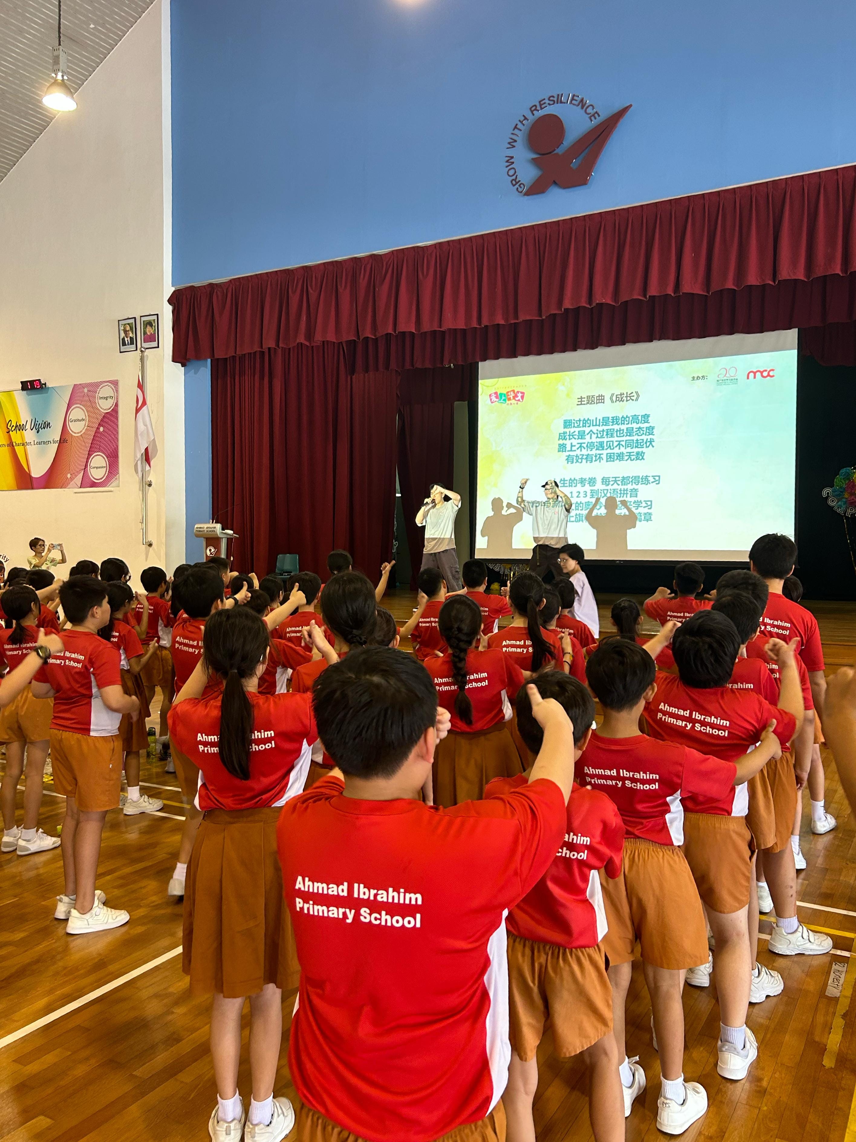 Students in red Ahmad Ibrahim Primary School shirts face a stage with a presenter and a Chinese-text projection screen.