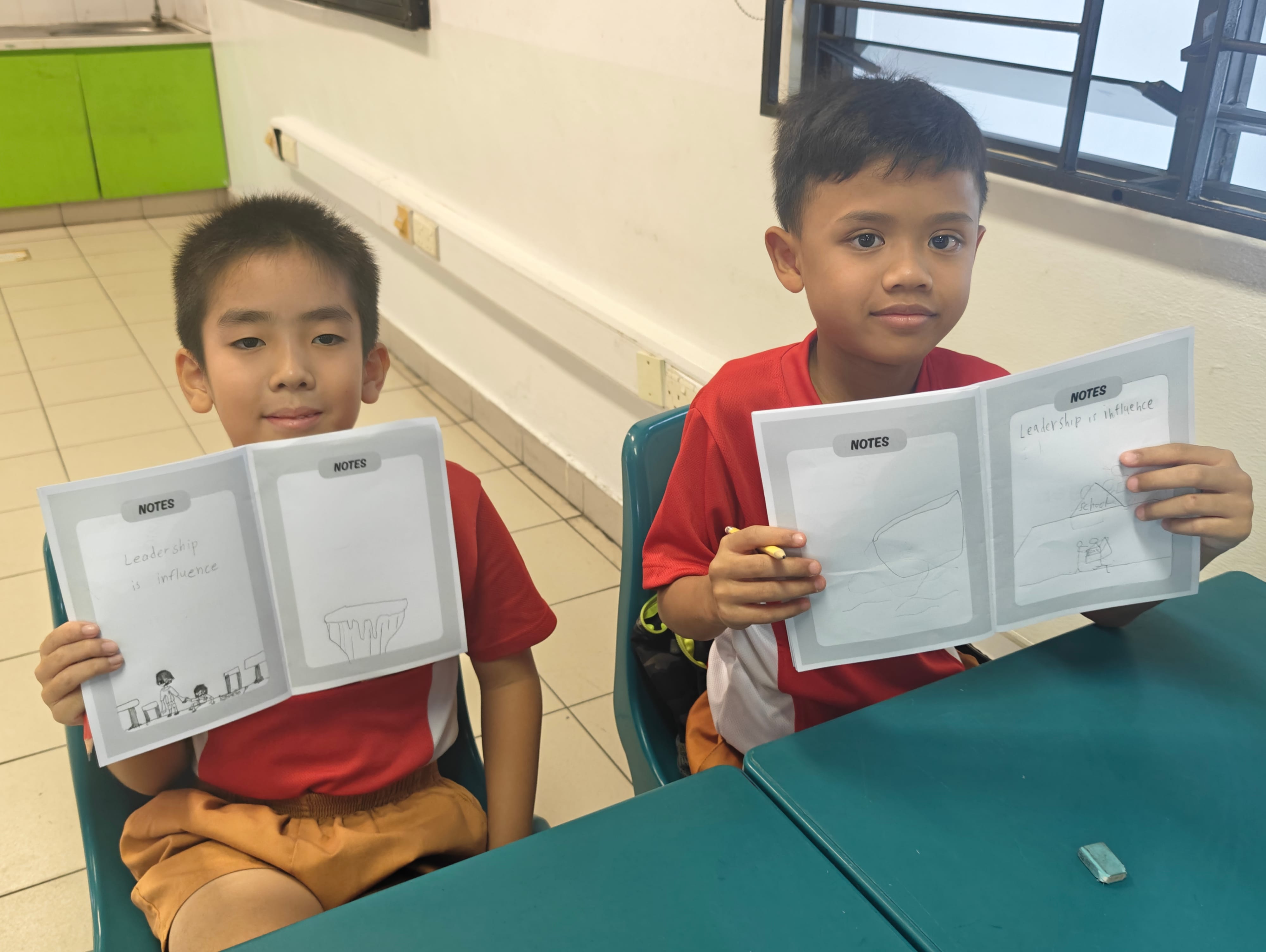 Two boys in red shirts sit at a desk, each holding a notes booklet titled "Leadership is Influence."