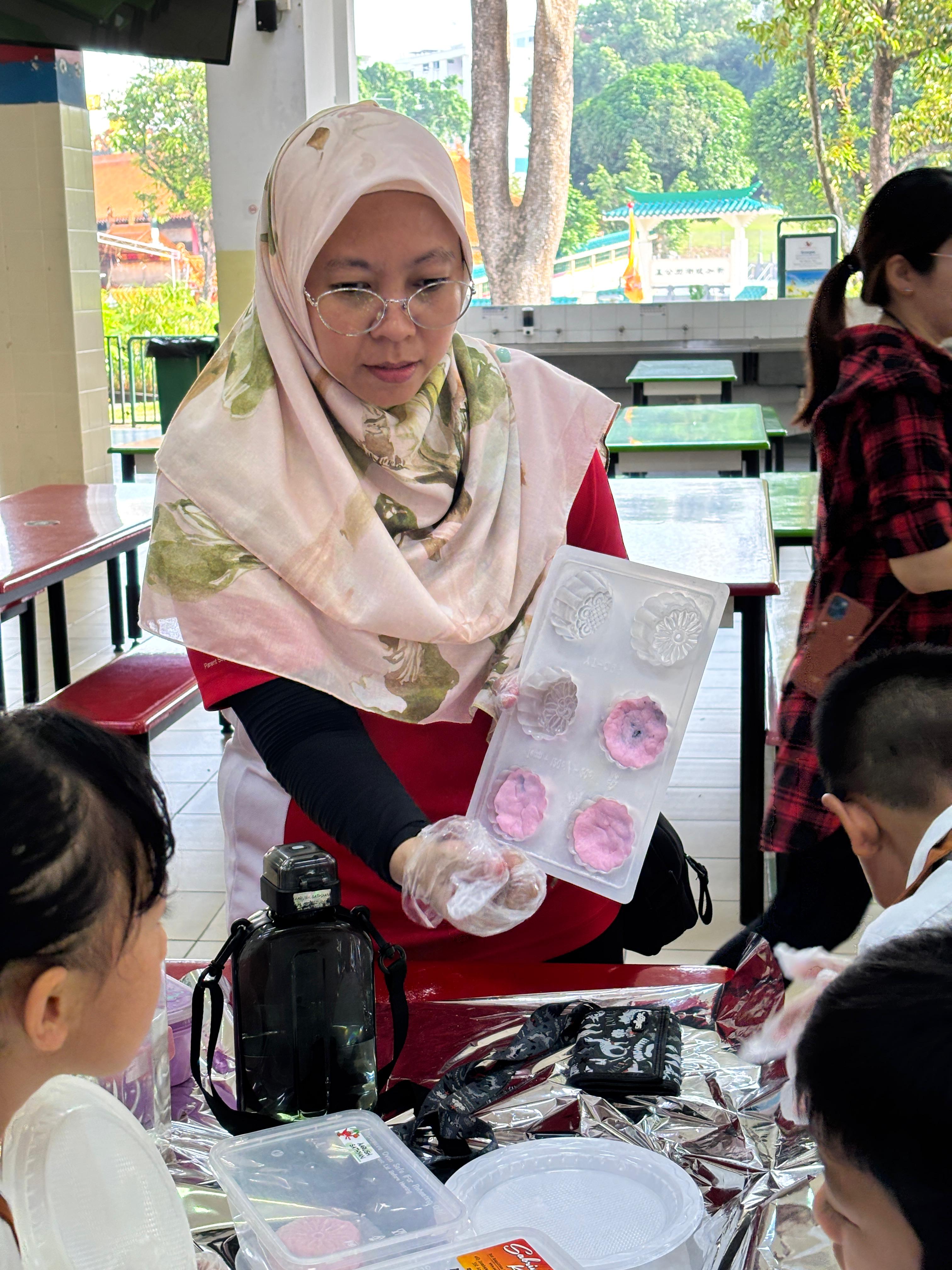 Woman in hijab and glasses holds a mooncake mold tray with pink mooncakes while children watch at a table.