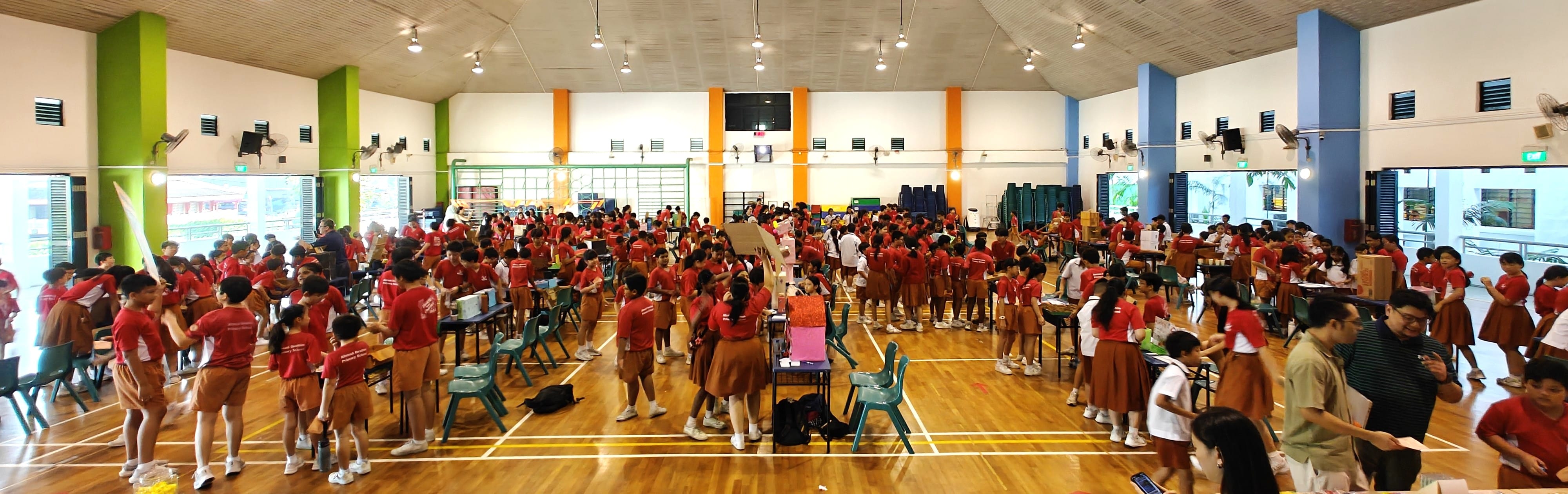 Large school hall filled with students in red tops and brown uniforms gathered around tables during an indoor activity.