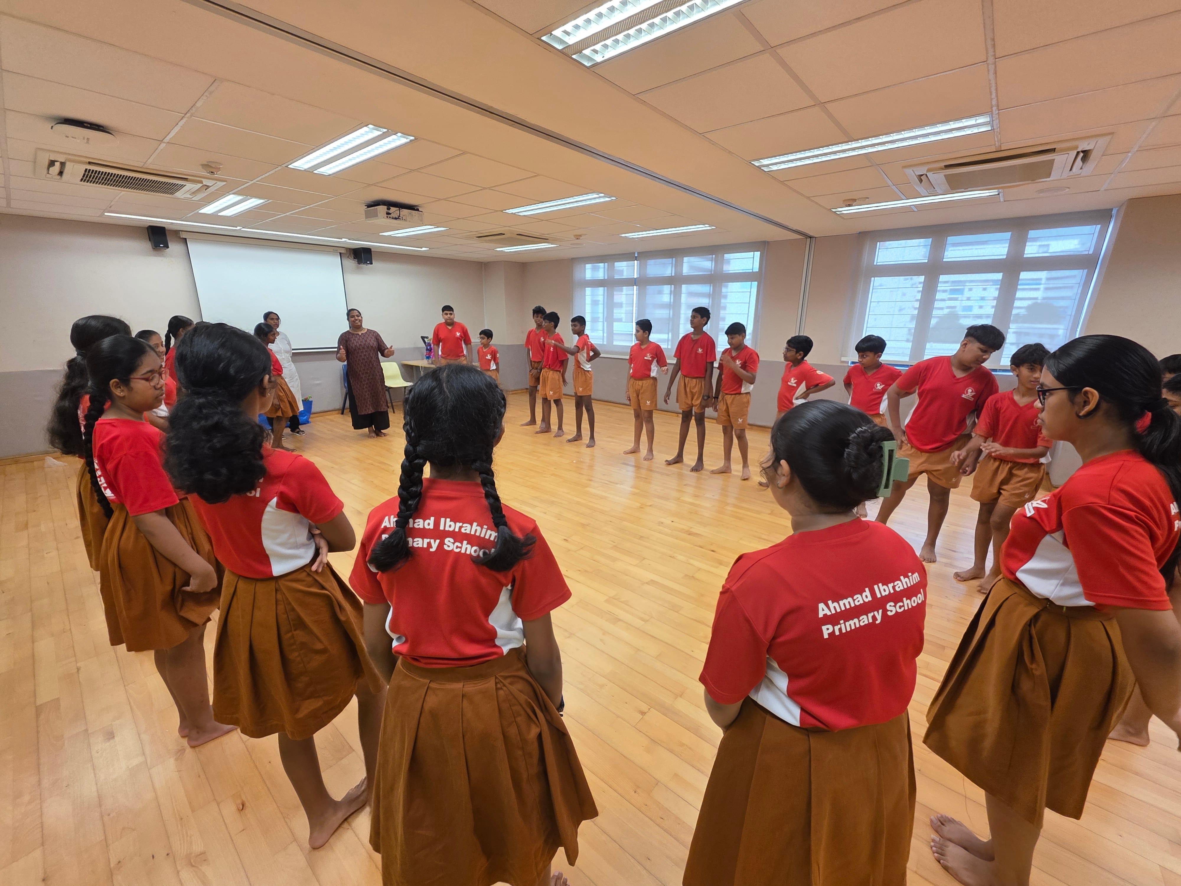 Children in Ahmad Ibrahim Primary School uniforms stand in a circle on a wooden floor during a dance class.