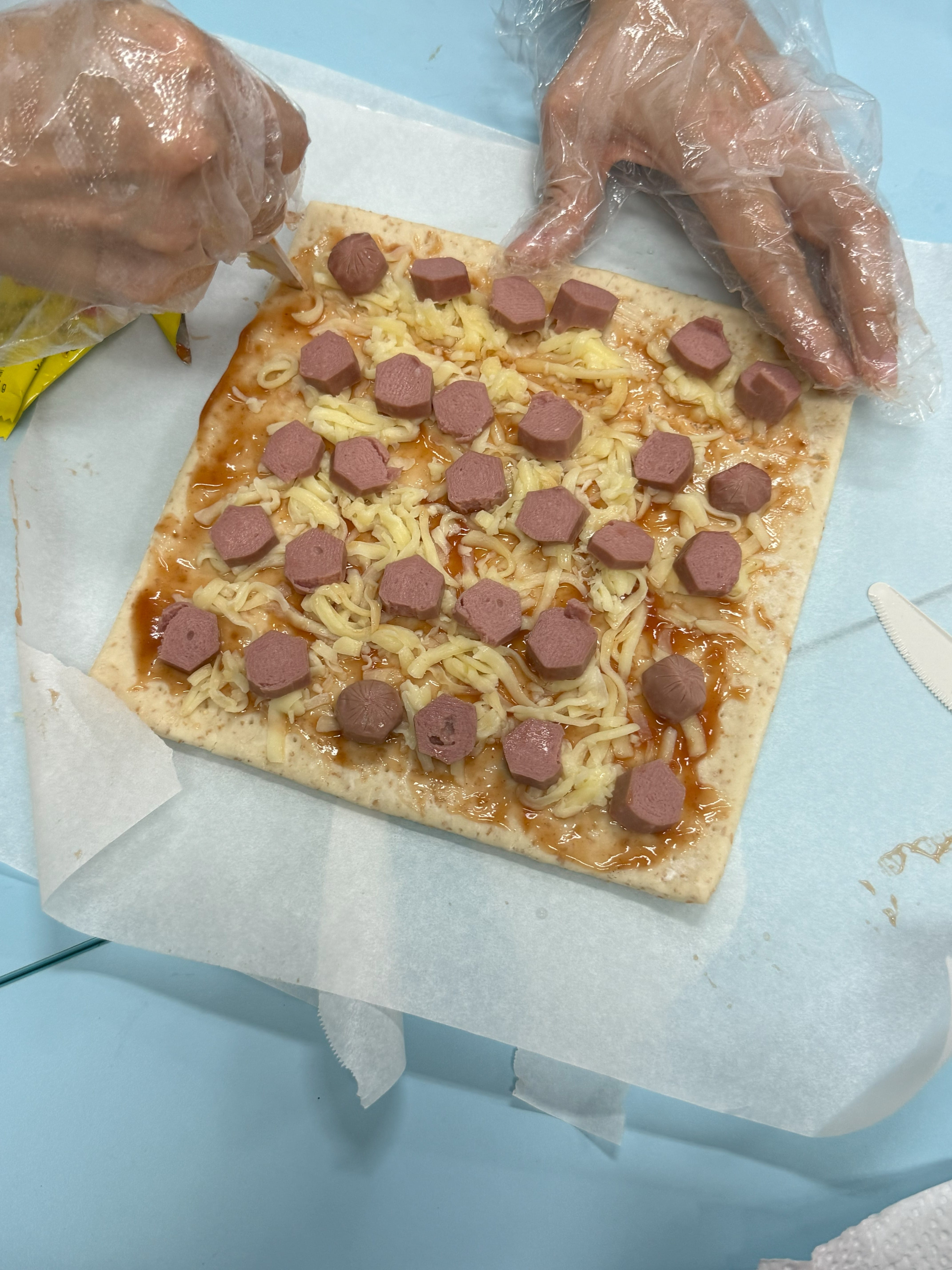 Gloved hands arranging sliced sausage pieces on a square unbaked pizza with shredded cheese and sauce.