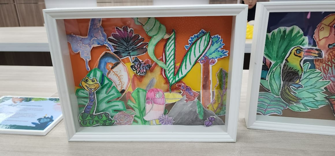 Two framed children's artworks showing colorful jungle scenes with frogs, plants, butterflies, and a toucan.