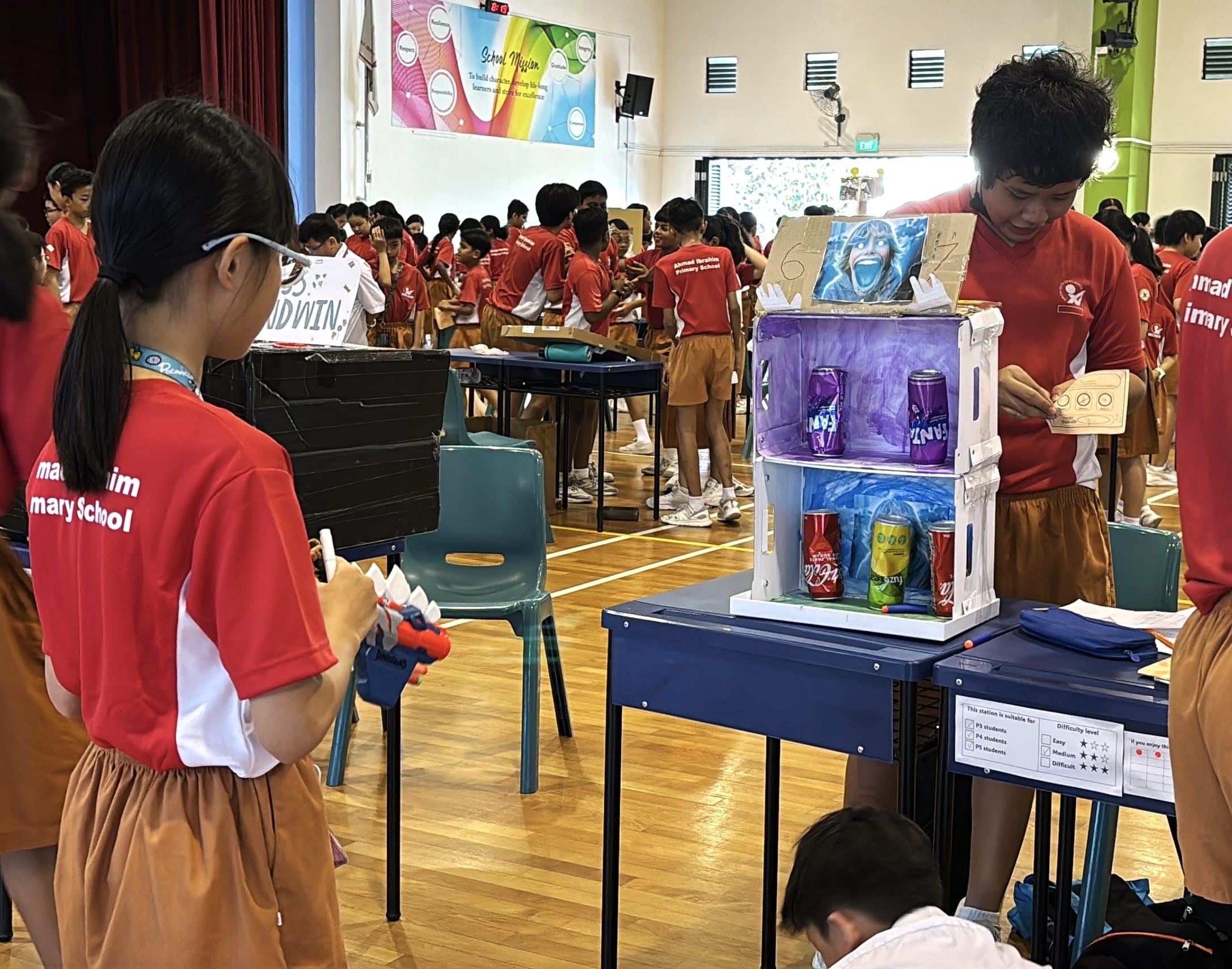Students in red uniforms gathered in a school hall around a homemade cardboard vending machine holding colorful cans.