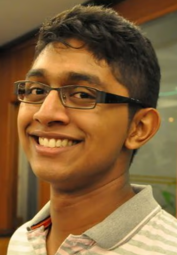 Smiling person with short dark hair and black rectangular glasses, wearing a light striped collared shirt.