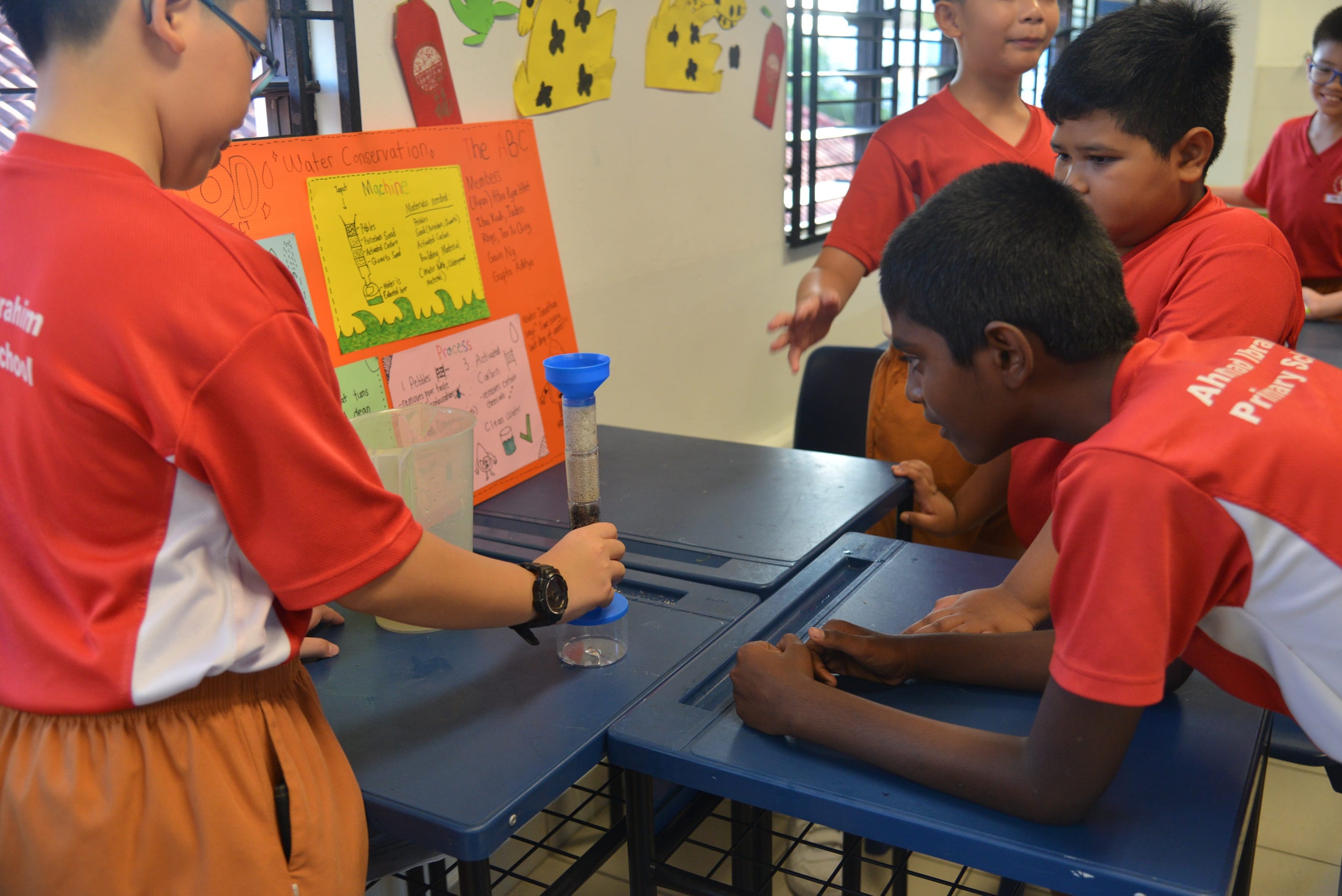 Students in red uniforms gather around a desk with a water conservation science fair project display board.