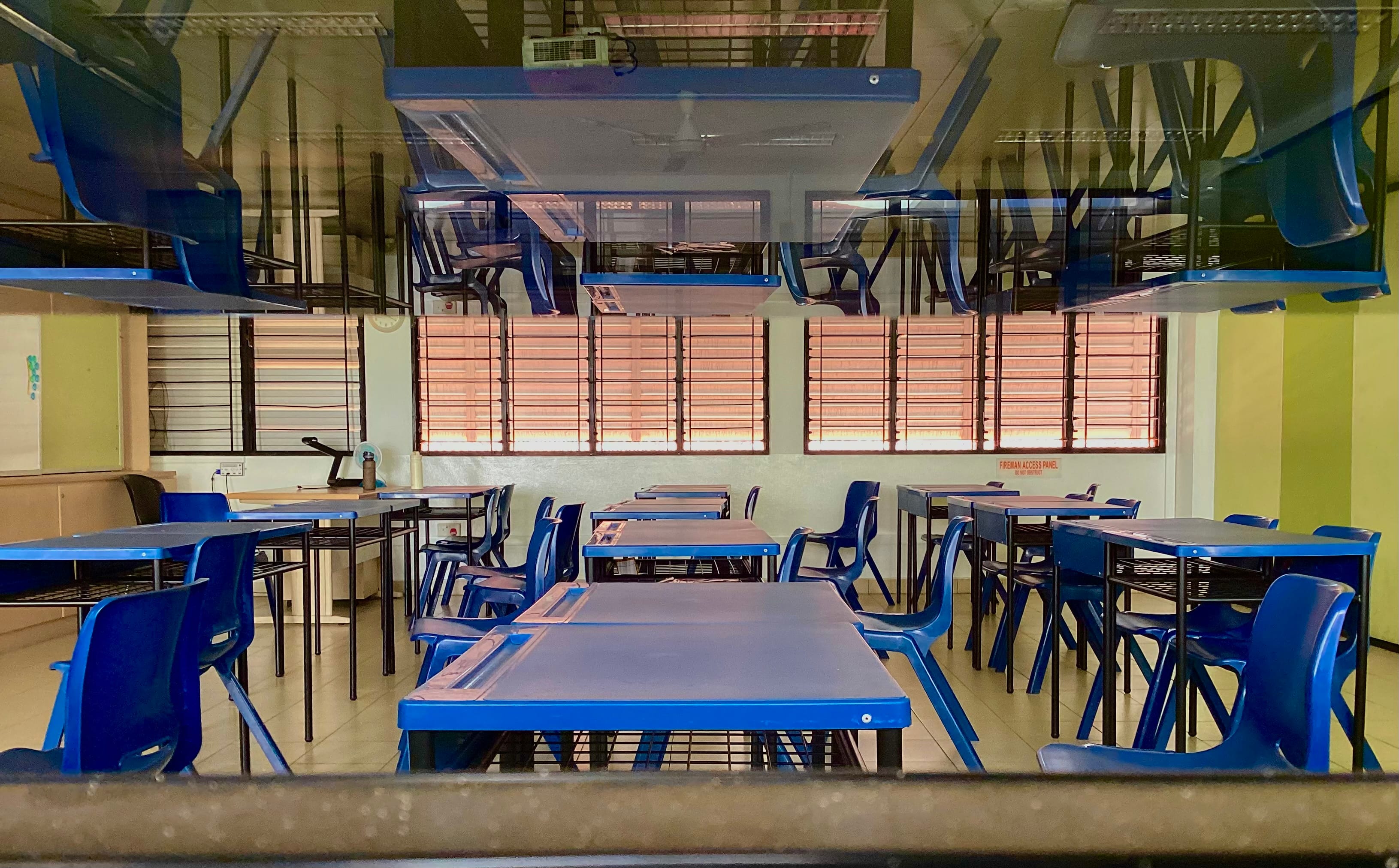 Empty classroom with rows of blue chairs and desks, viewed through a glass window reflecting the furniture above.