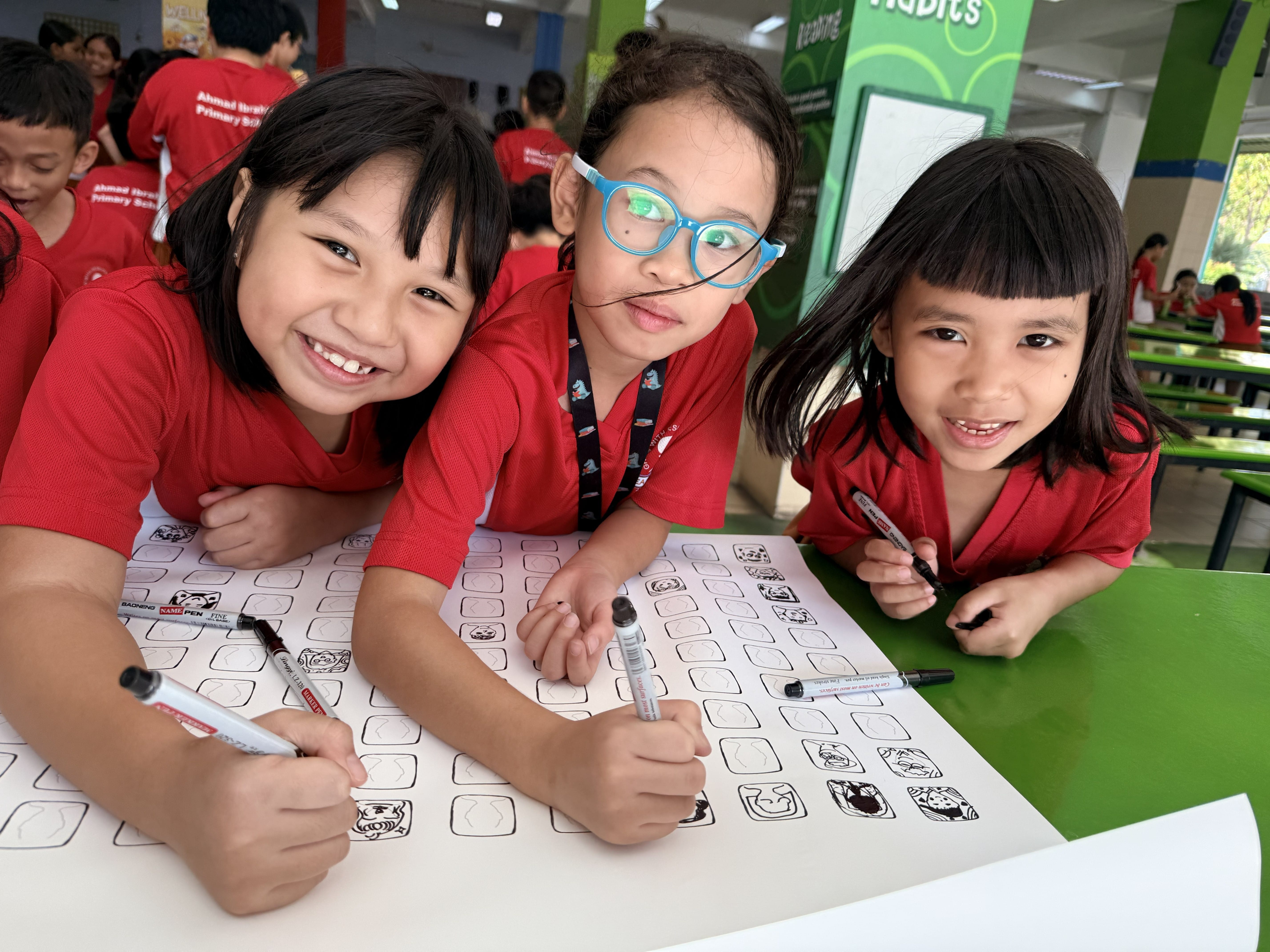 Three smiling girls in red school uniforms lean over a sheet of drawings, holding markers in a school hall.