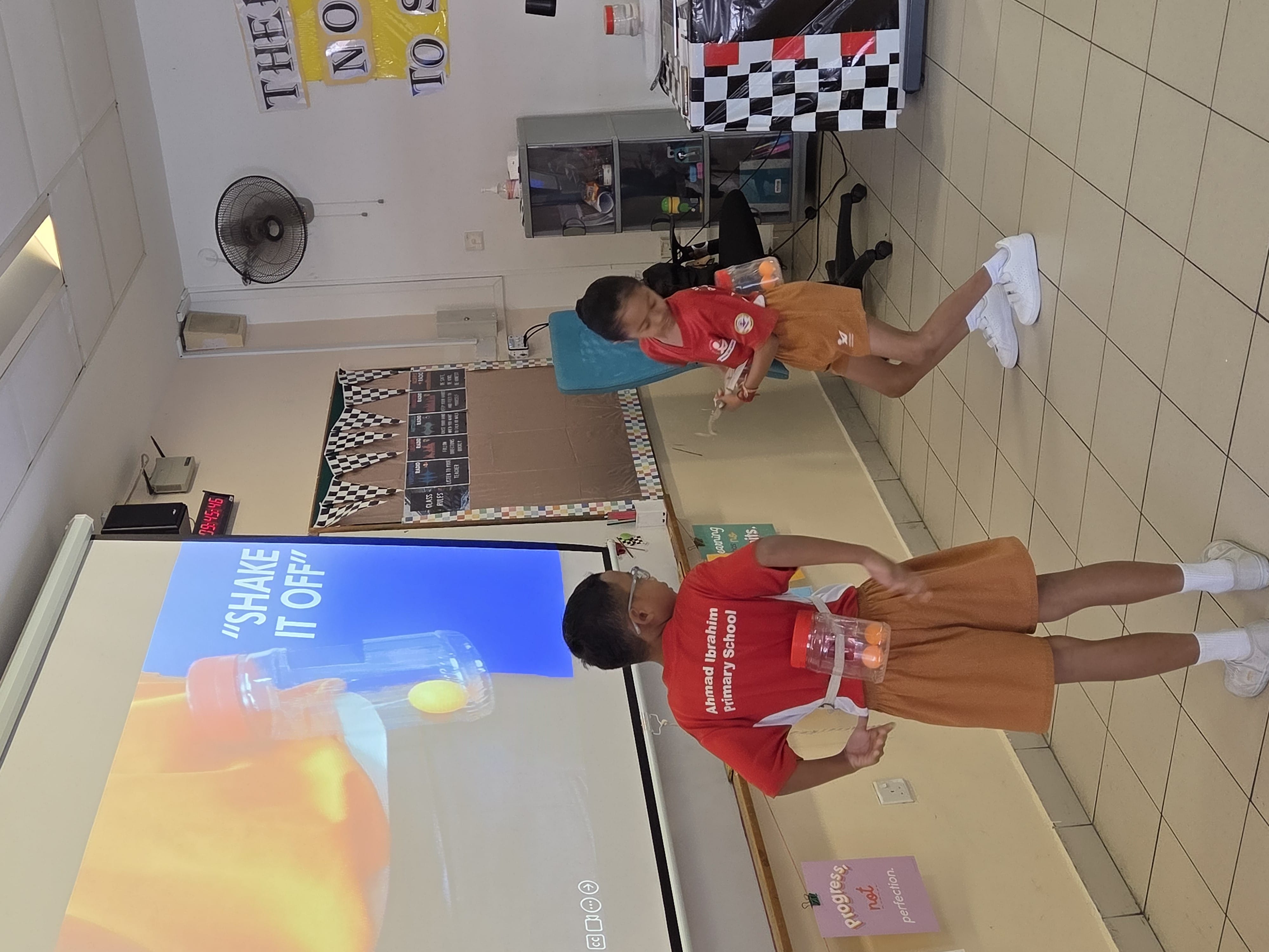 Two children in red school uniforms shake bottles strapped to their backs in a classroom with a "Shake It Off" slideshow.