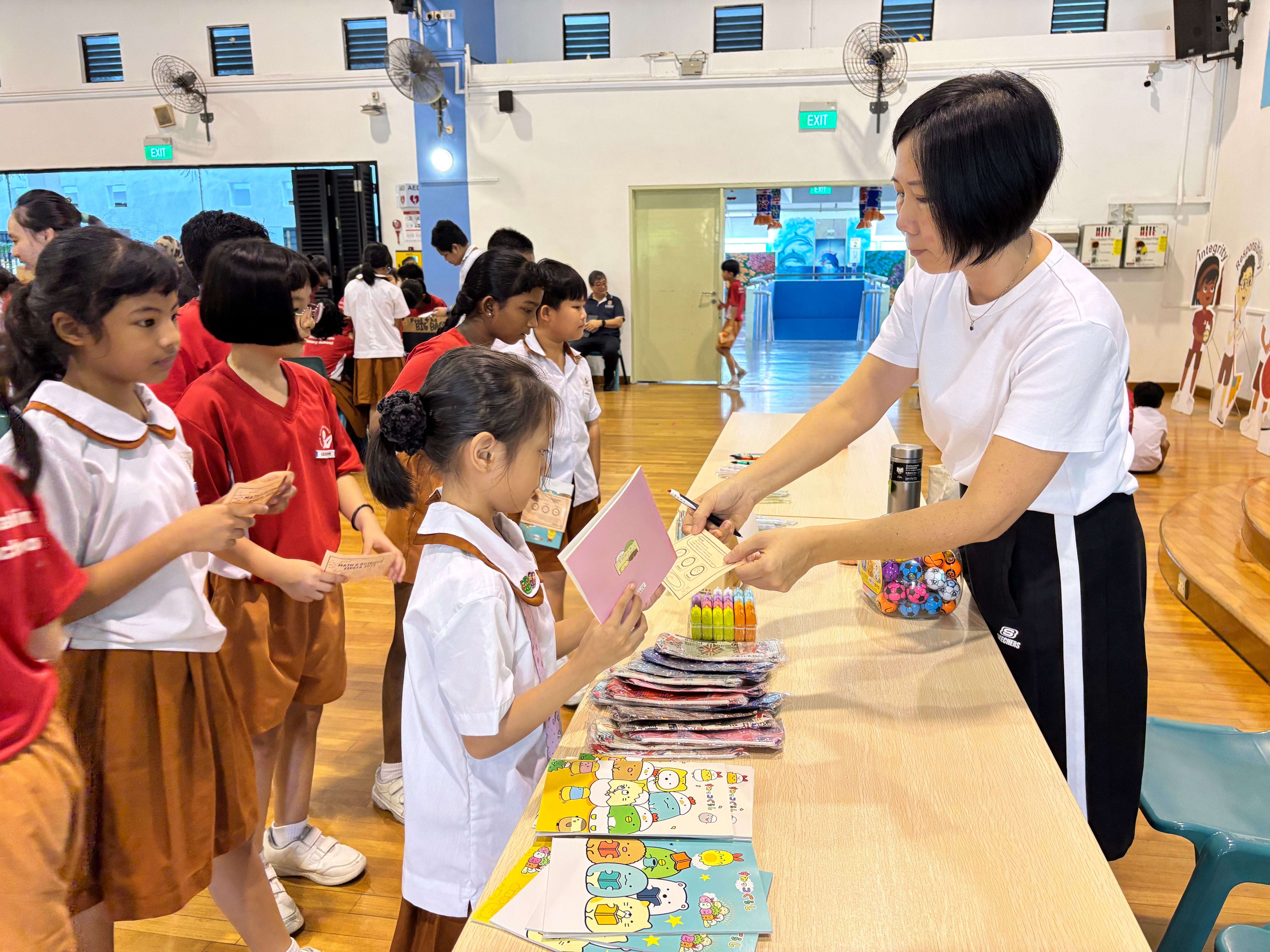 A woman in white hands out stationery to uniformed primary school children queuing at a table in a school hall.
