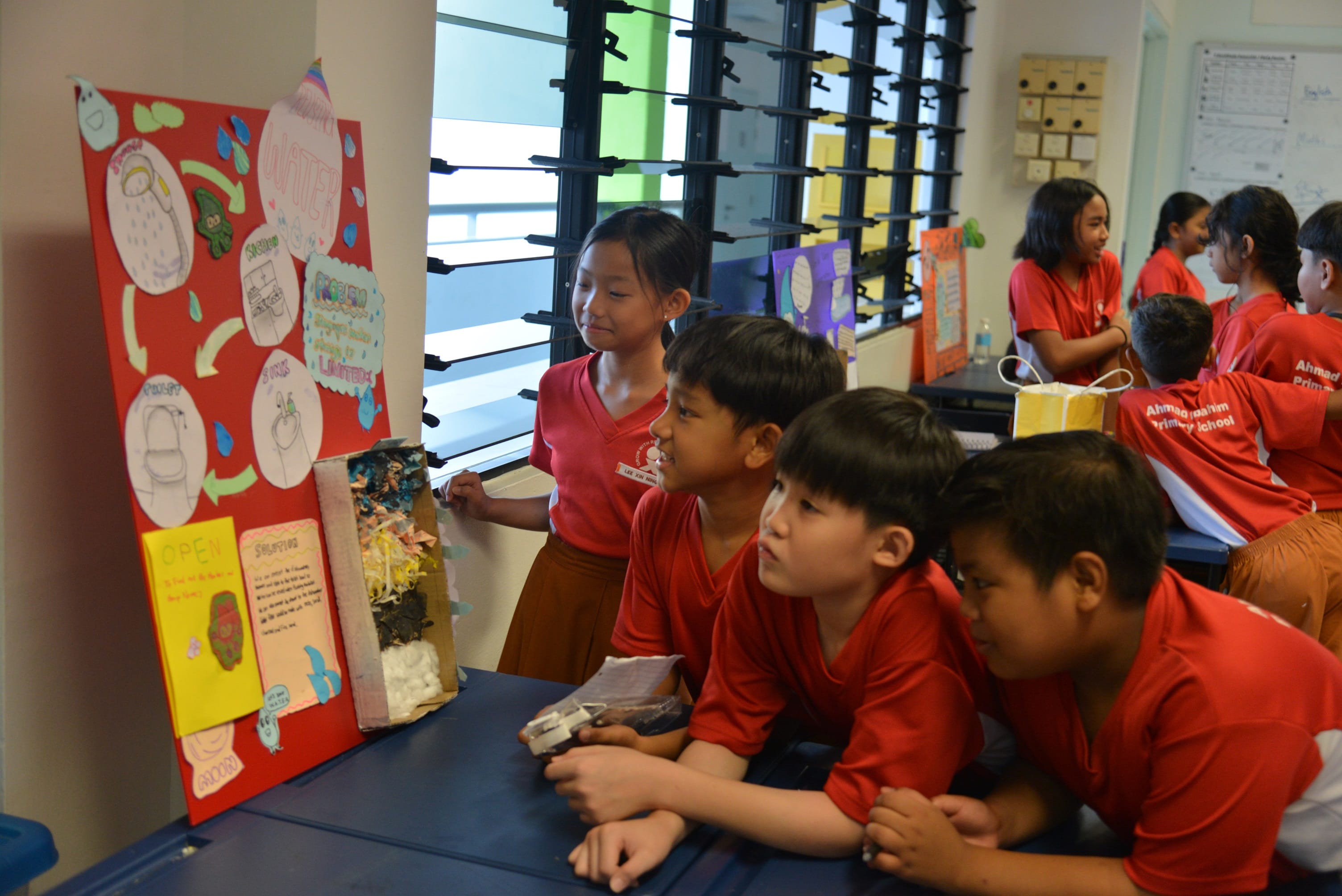 Primary school children in red uniforms look at a colorful red poster board project about water in a classroom.