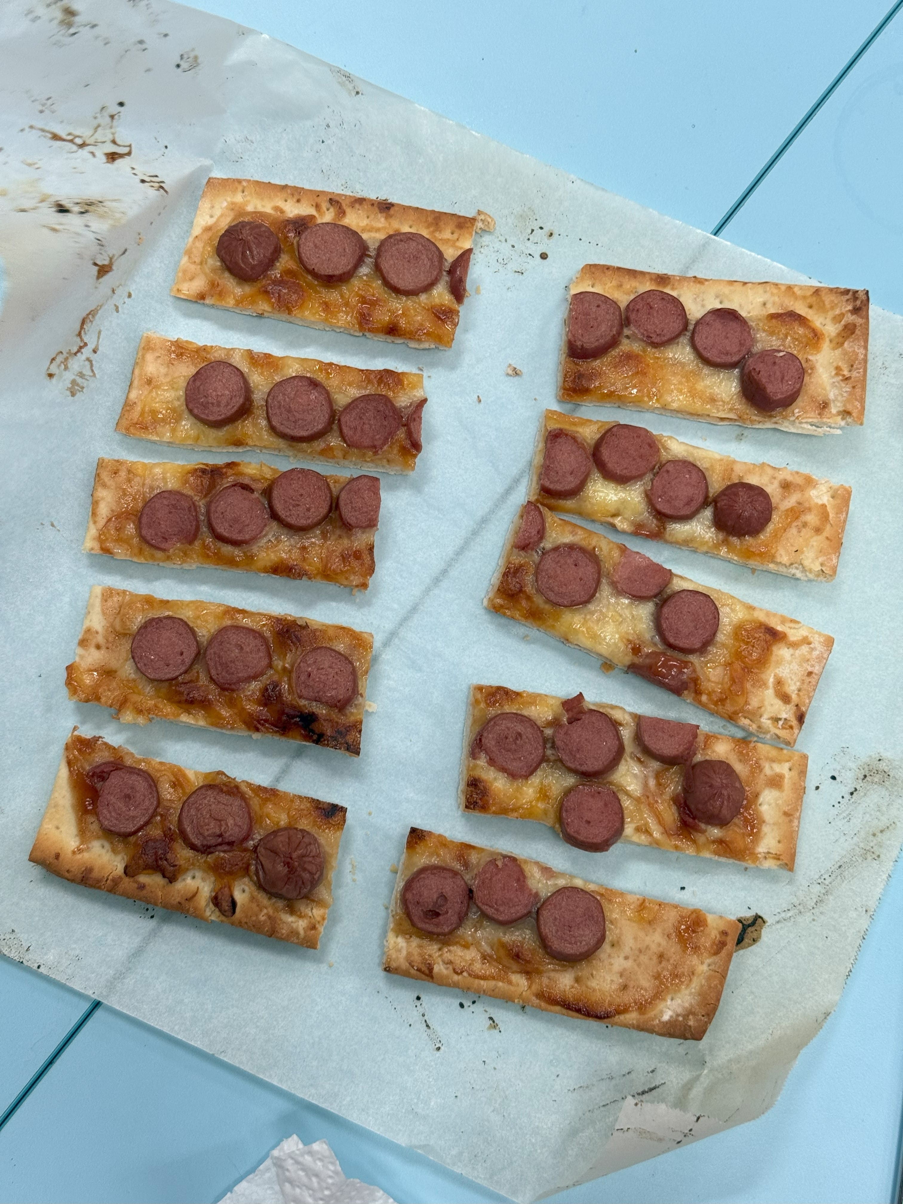 Ten rectangular flatbread pizzas topped with melted cheese and sliced sausage rounds on parchment paper.