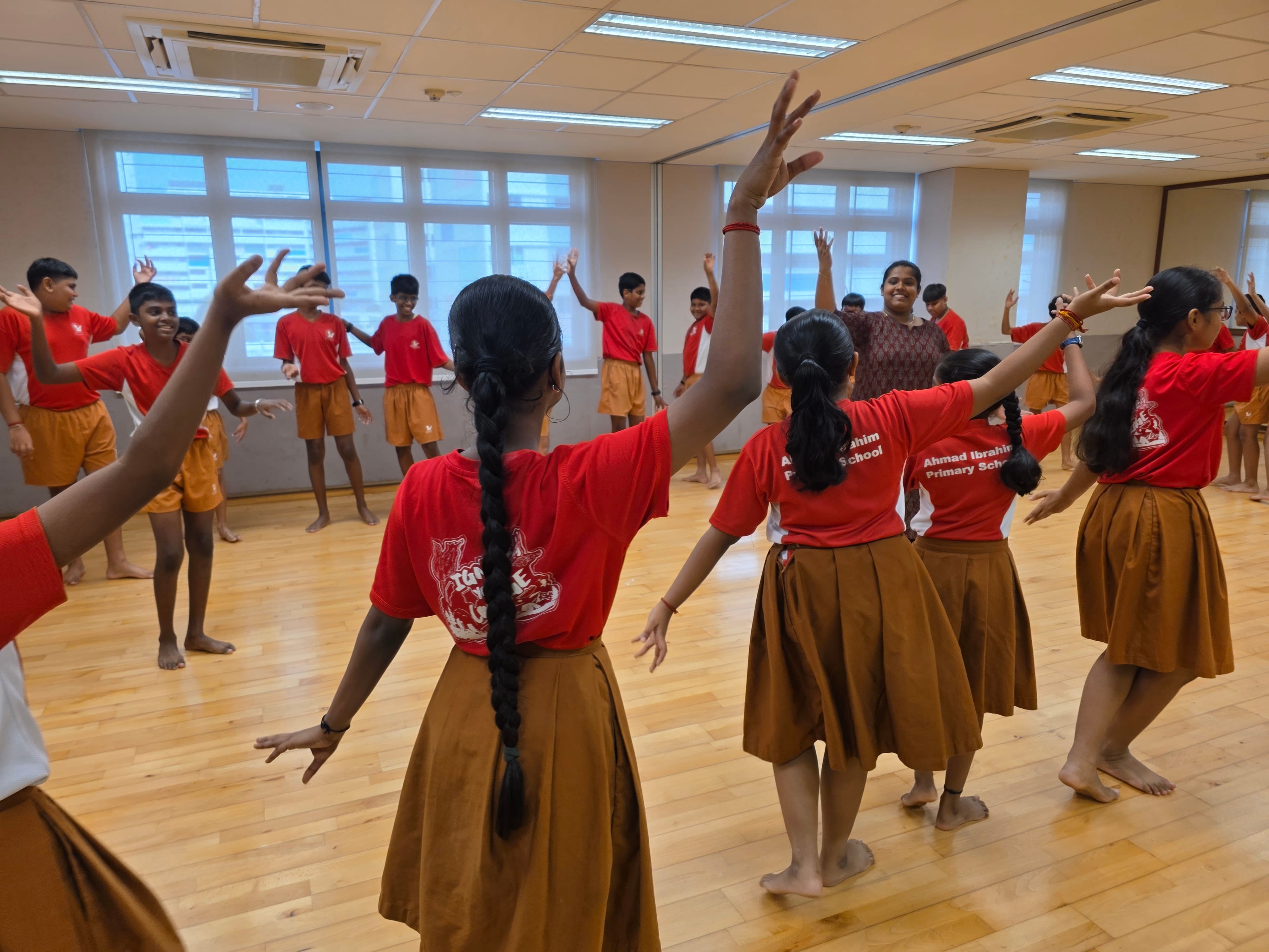 Children in red Ahmad Ibrahim Primary School uniforms practice a dance with raised arms in a studio.