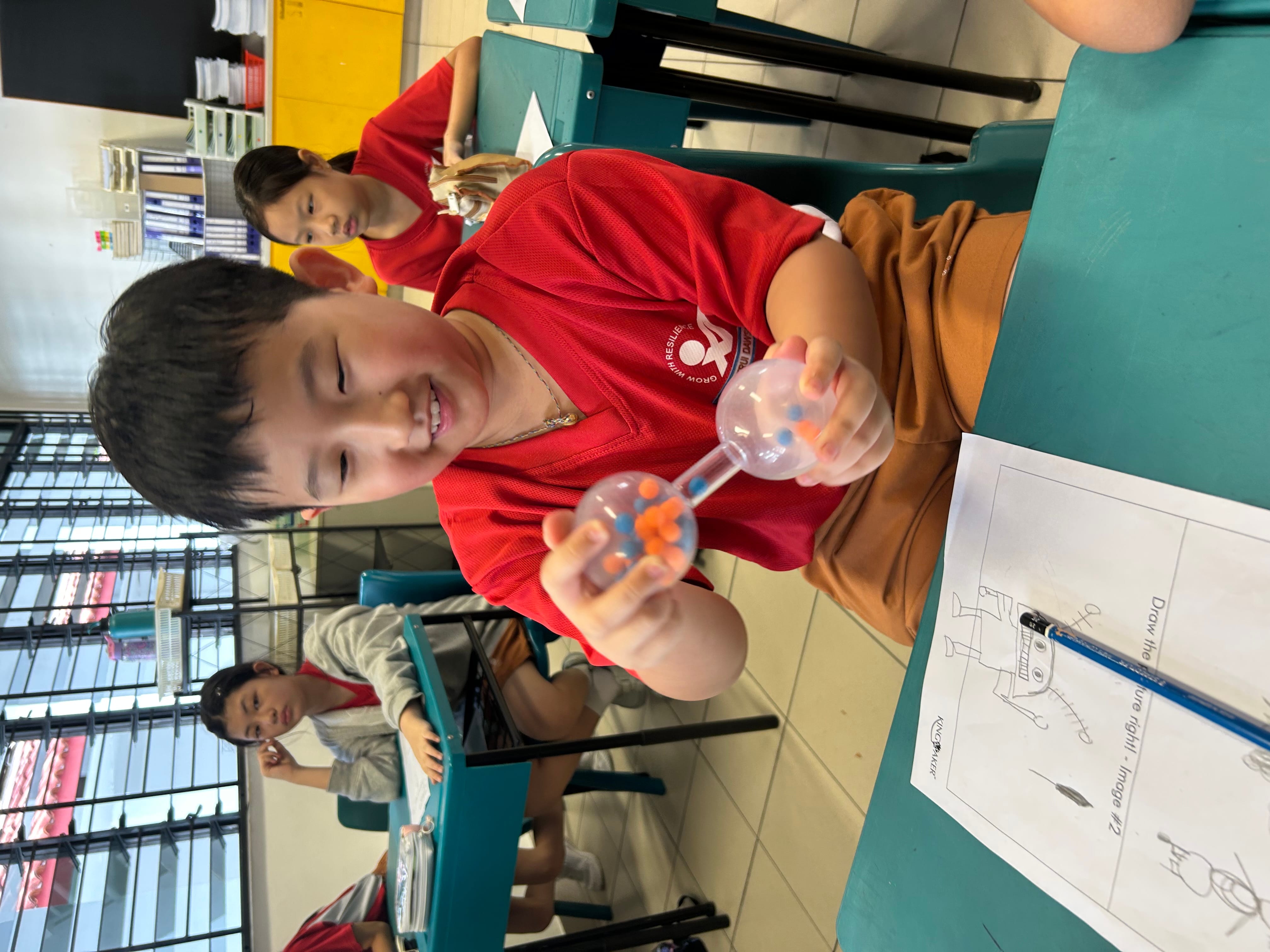 A smiling boy in a red shirt holds a clear hourglass toy with colorful beads in a classroom.