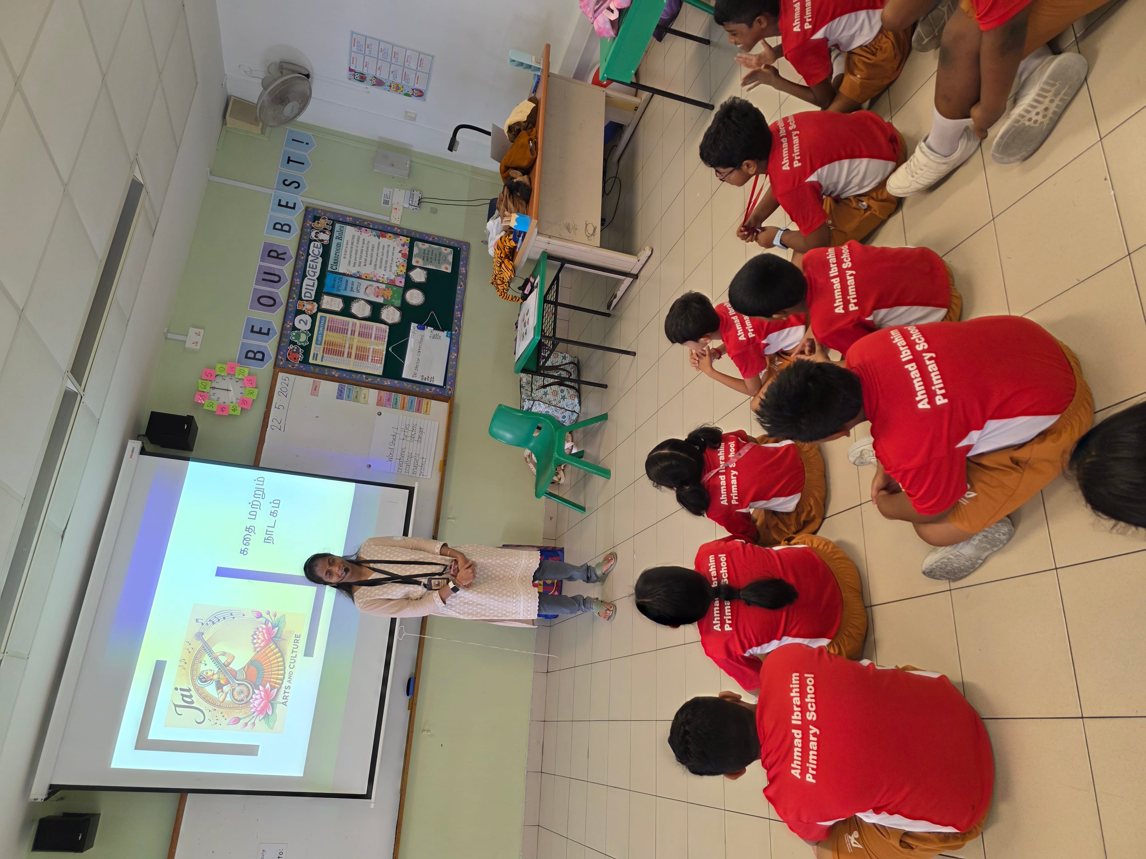 A teacher stands before a projected slide as students in red Ahmad Ibrahim Primary School uniforms sit on the floor.