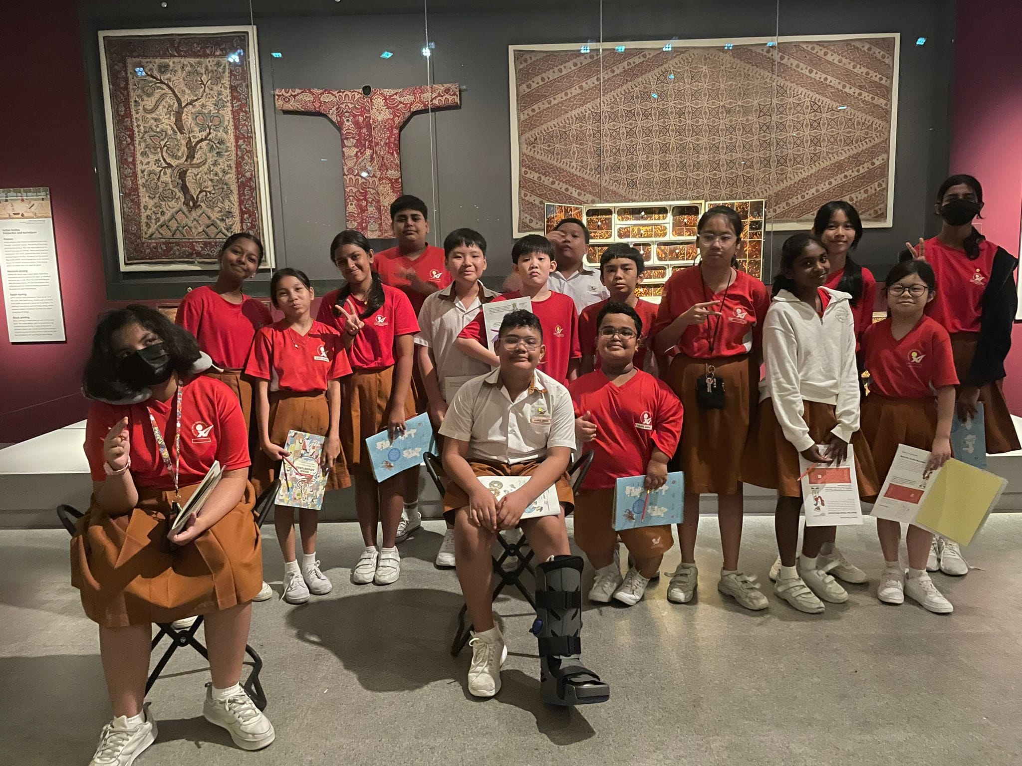 A group of students in red uniforms poses in a museum gallery with ornate textile displays behind them.