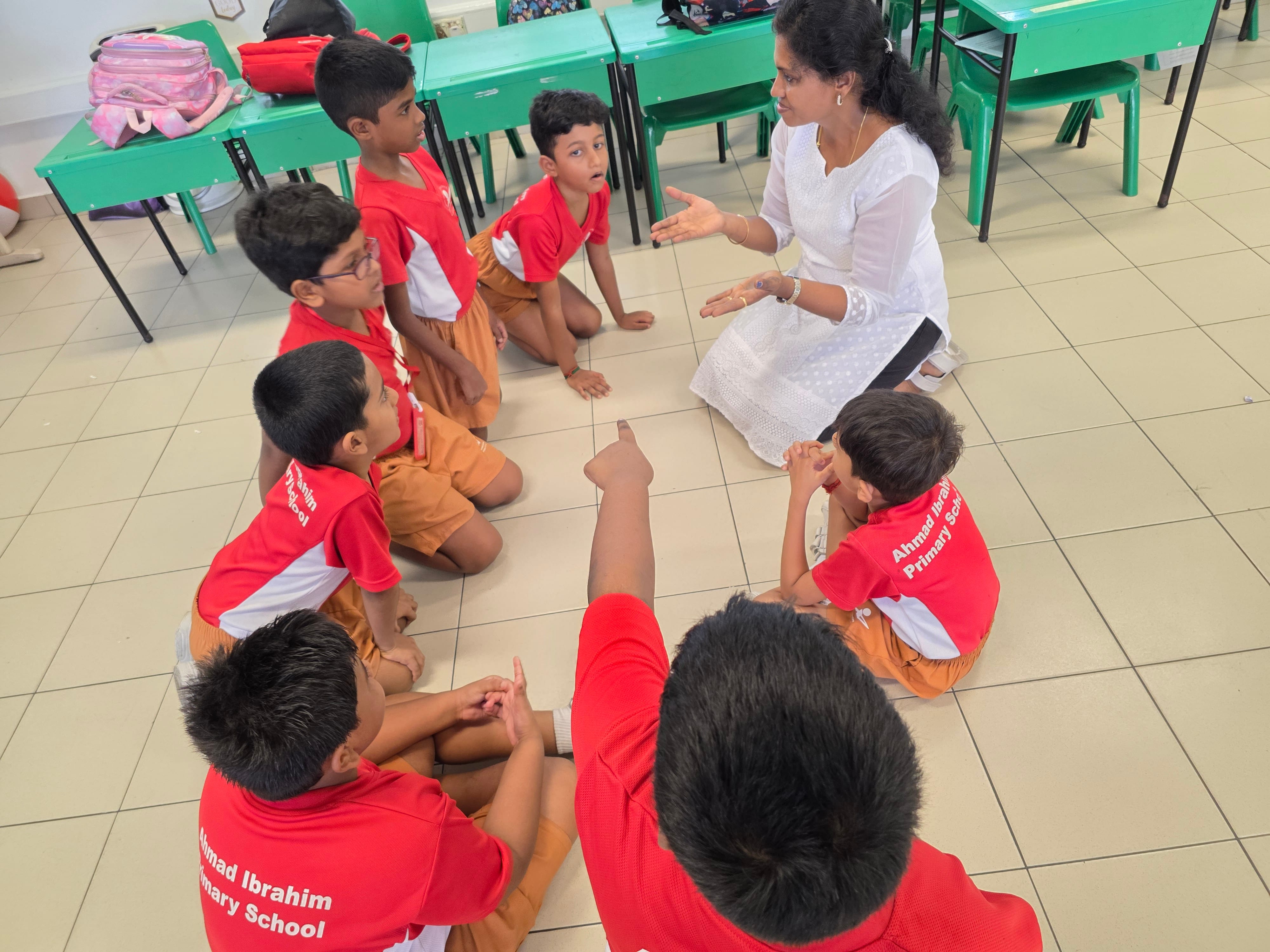 A woman in white sits on a classroom floor speaking to young boys in red Ahmad Ibrahim Primary School uniforms.