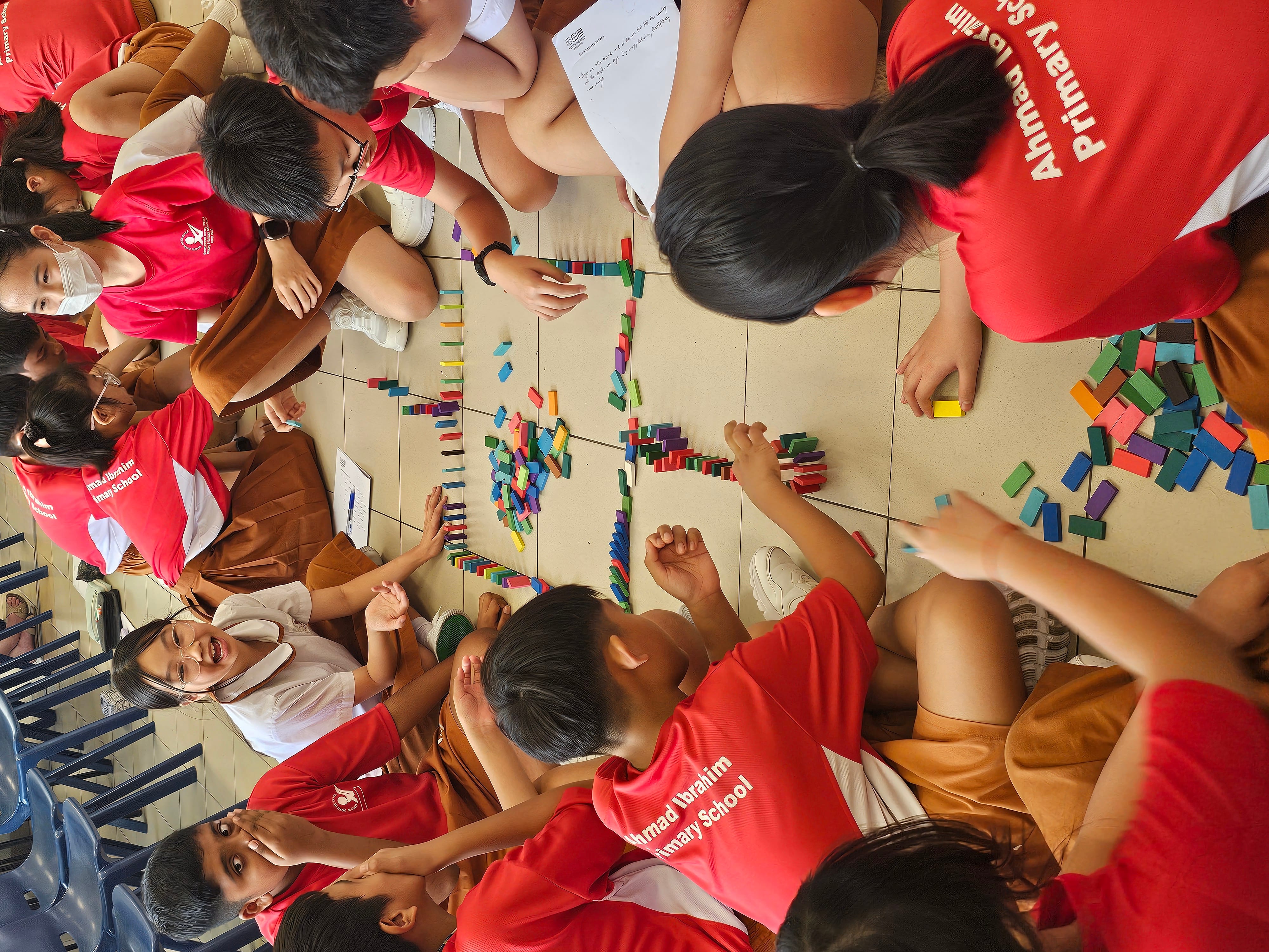 Students in red Ahmad Ibrahim Primary School shirts sit on a floor arranging colorful dominoes in a pattern.
