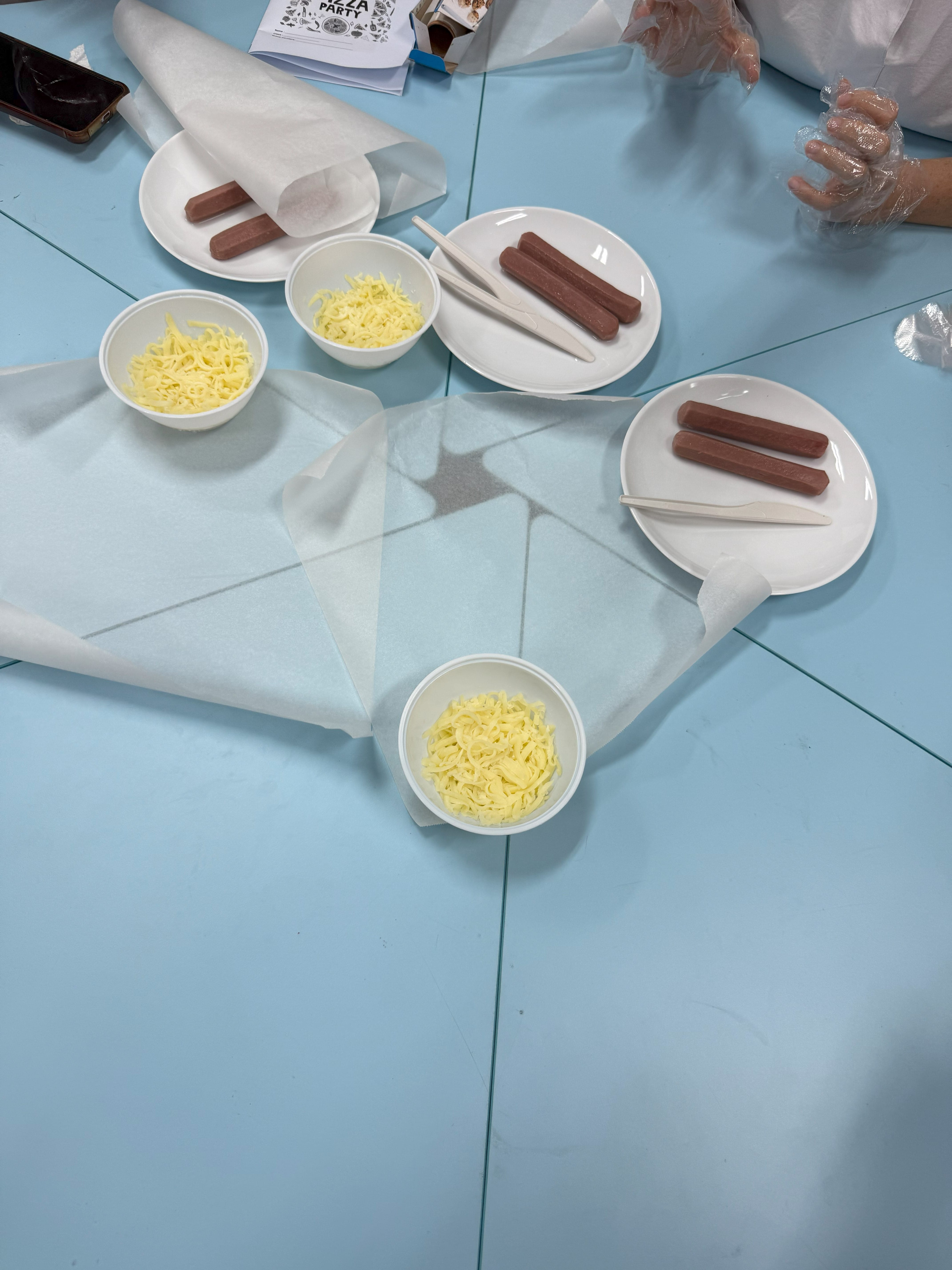 White plates with sausages and bowls of shredded cheese on a blue table with parchment paper.
