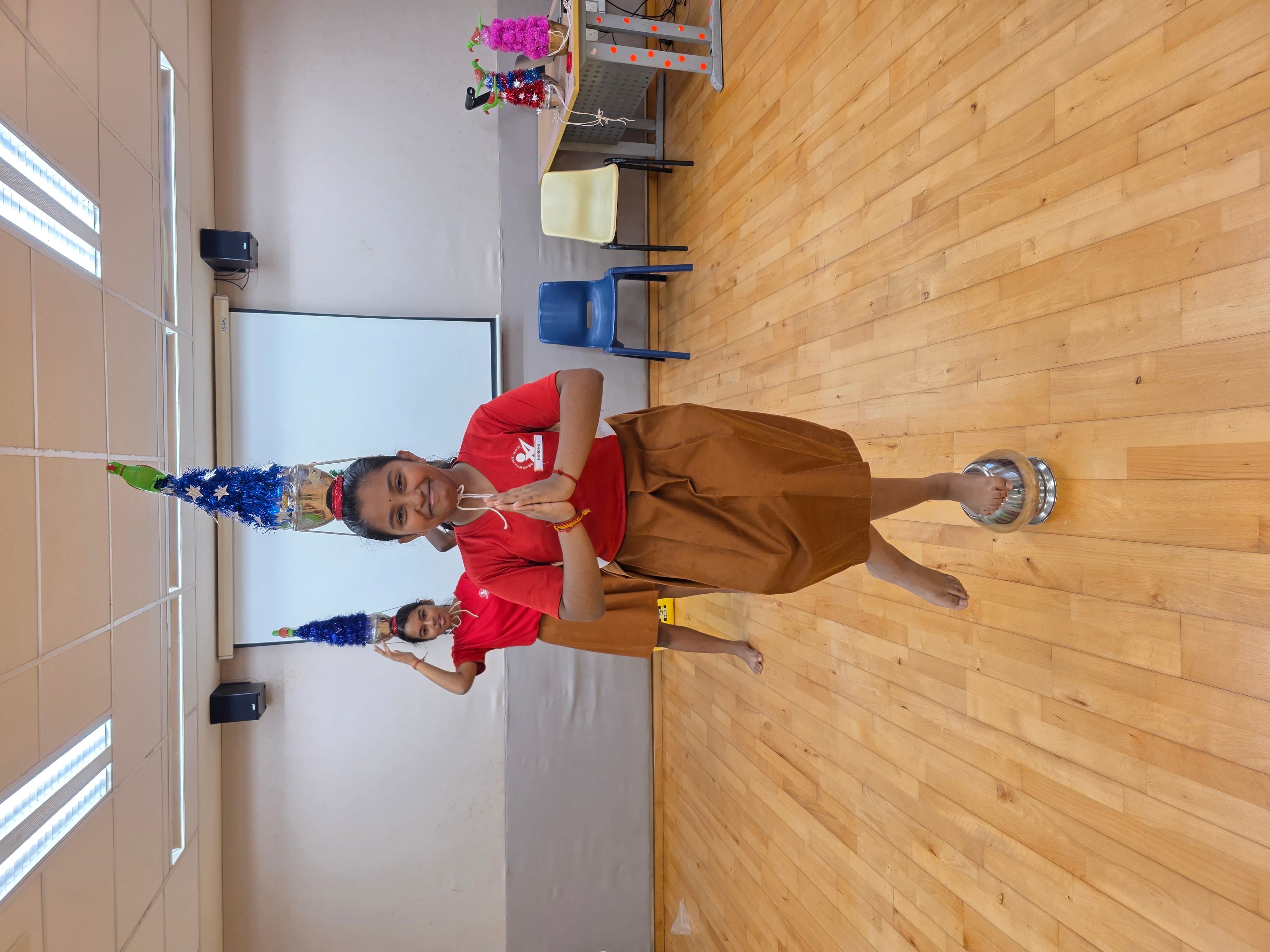 Girl in red top balances decorated pot on head and stands on a metal bowl, hands in prayer pose, in a school hall.