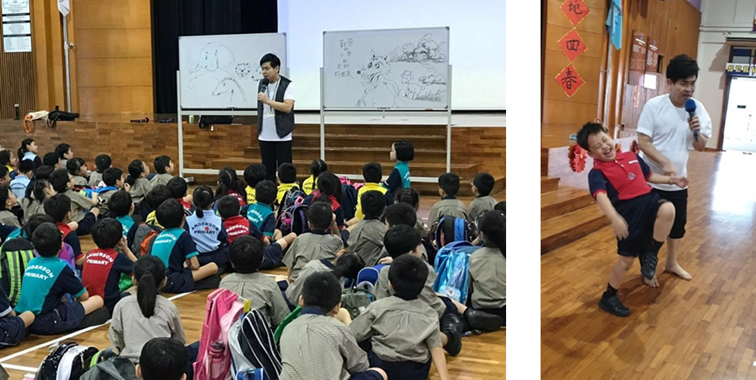 Students seated on gym floor watching a man with mic by a whiteboard; at right he holds a laughing boy.