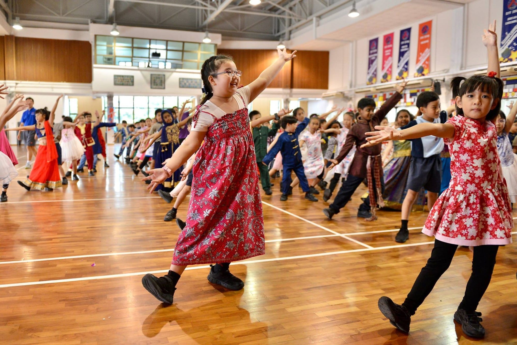 Children in colorful cultural outfits dancing together in a school gymnasium with arms outstretched.