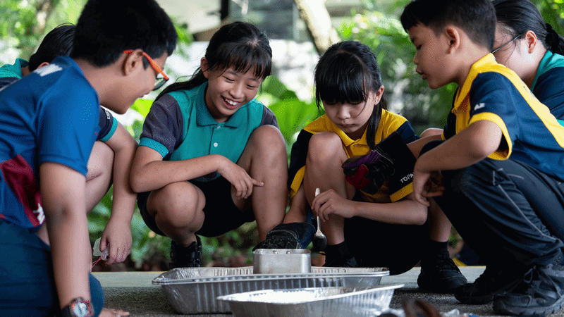 Four children crouch outdoors around metal trays on the ground, smiling and engaged in a hands-on activity.