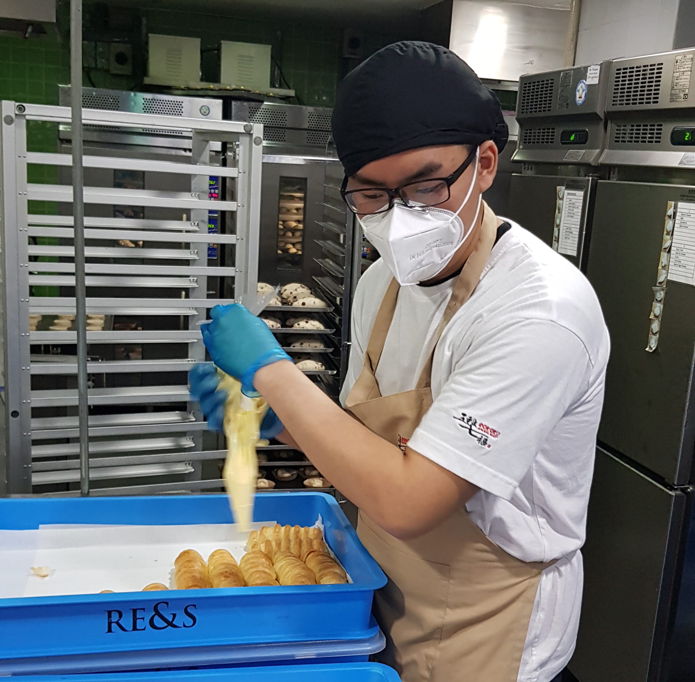 A baker wearing a mask, gloves, and apron handles dough over a blue tray of pastries in a commercial kitchen.