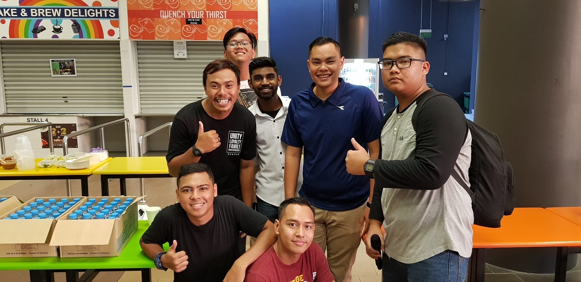 A group of seven men posing together in a canteen near food stalls, several giving a thumbs up, with drink crates on a table