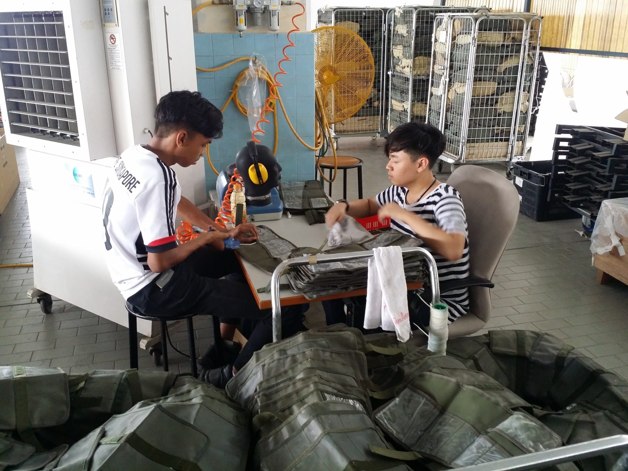 Two young men sit at a workbench in a workshop, handling olive green military-style bags and gear.